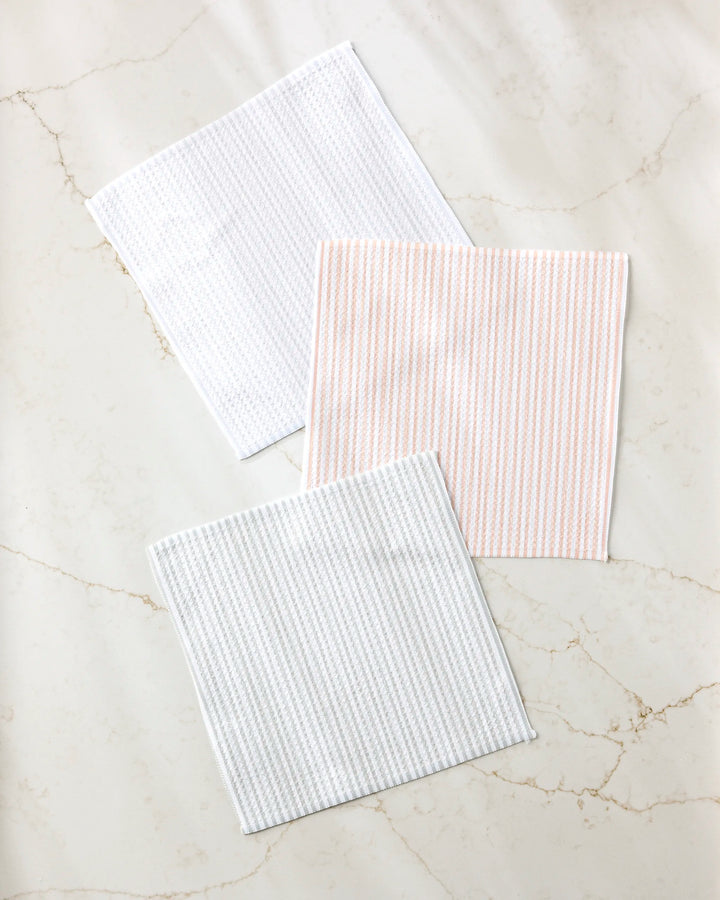 Three square À La Carte cloth napkins by Kailee Wright, made from recycled materials, are displayed on a light marble surface: one plain white, one with light pink stripes, and one with light gray stripes.