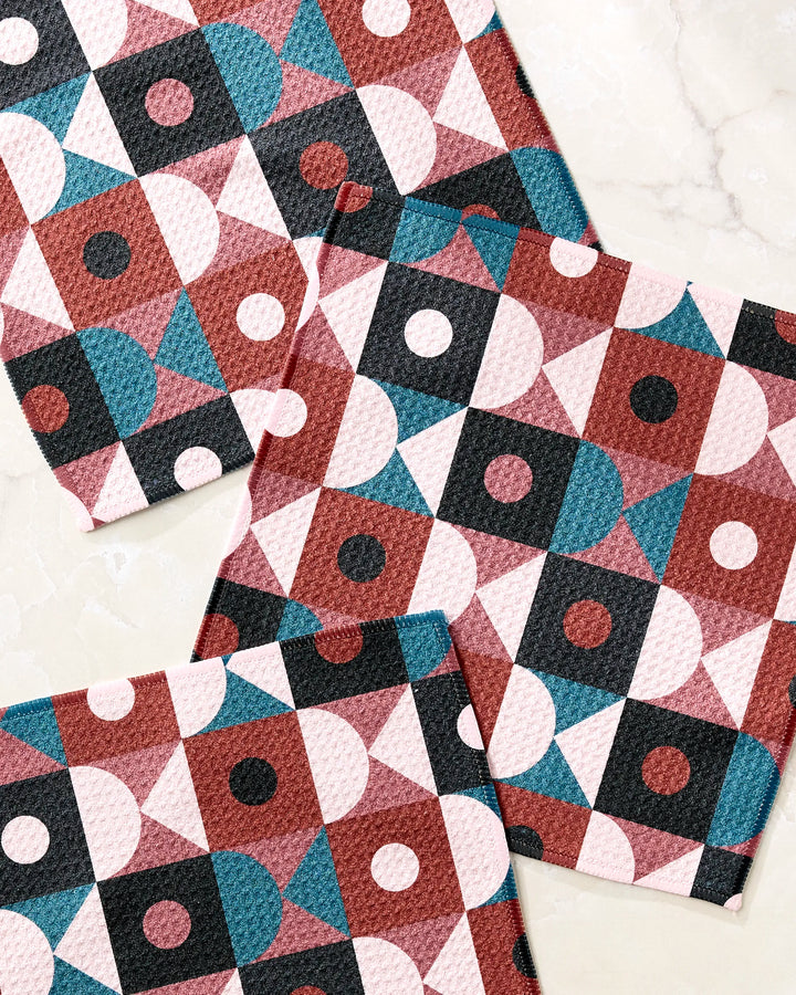 The Abstract Retro dishcloth set by Jennifer Bouron includes three square cloths featuring a geometric pattern in pink, burgundy, teal, white, and black on a marble-like background. Made with recycled materials.