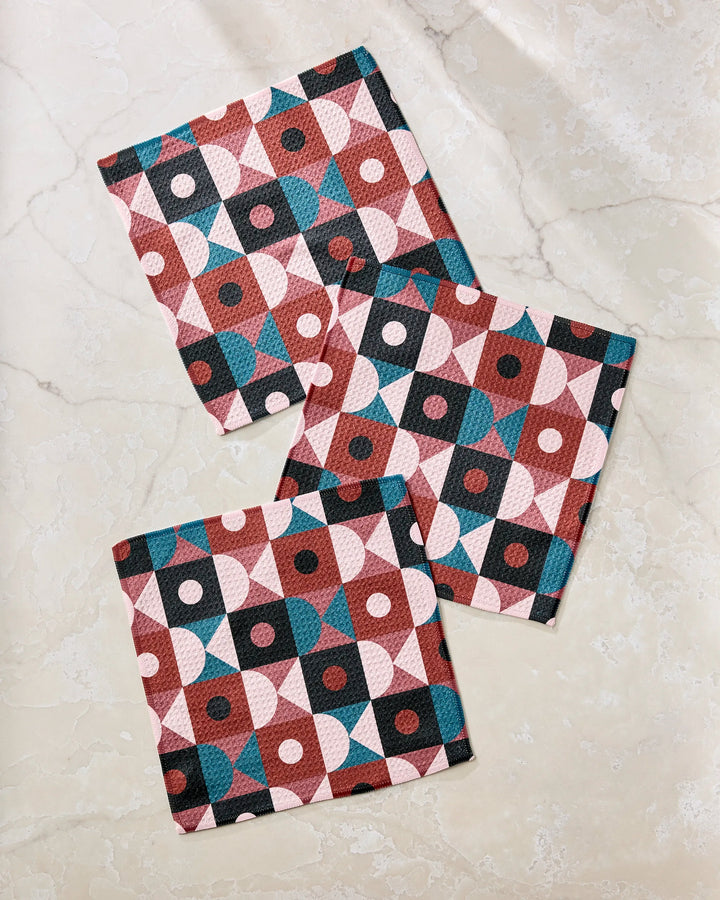 Three Abstract Retro napkins by Jennifer Bouron, made from recycled materials, display teal, maroon, pink, and black geometric shapes and dots, neatly arranged on a light marble surface.