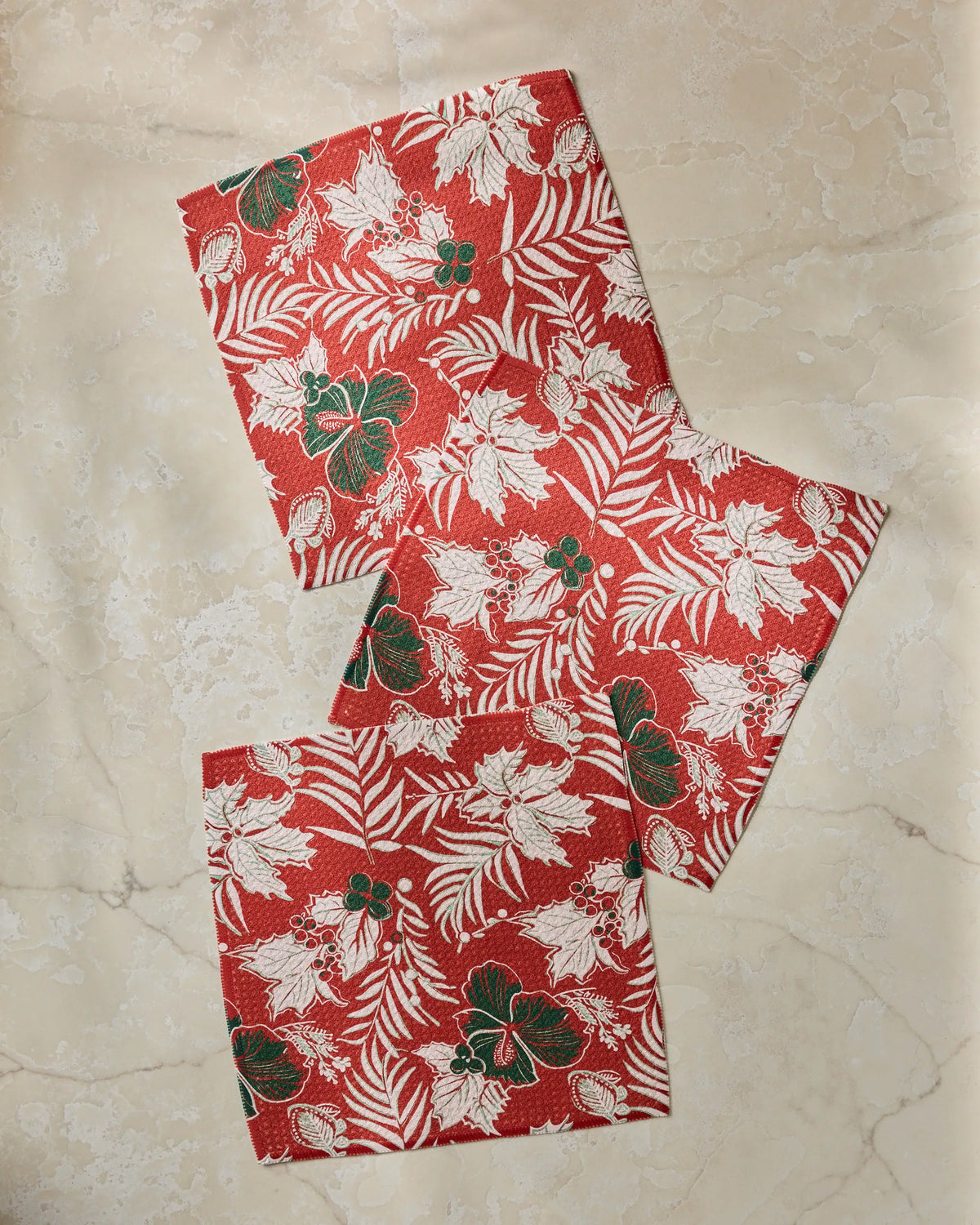 Three pieces from the Geometry Aloha Winters collection, featuring red fabric adorned with white and green tropical leaf and flower patterns, are displayed on a light marble surface—creating a vibrant dishcloth set crafted from recycled materials.