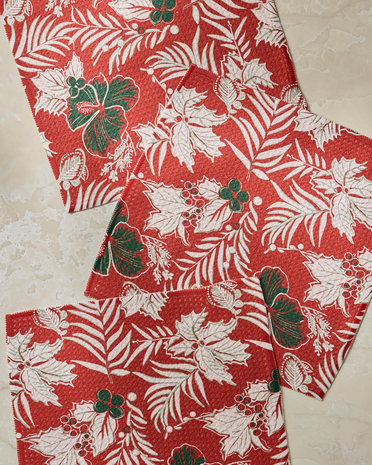 Three “Aloha Winters” napkins by Geometry, featuring red fabric adorned with white and green holiday-themed botanical patterns—leaves, berries, and ferns—are arranged on a light marble surface. This eco-friendly dishcloth set is crafted from recycled materials, perfect for your festive table.