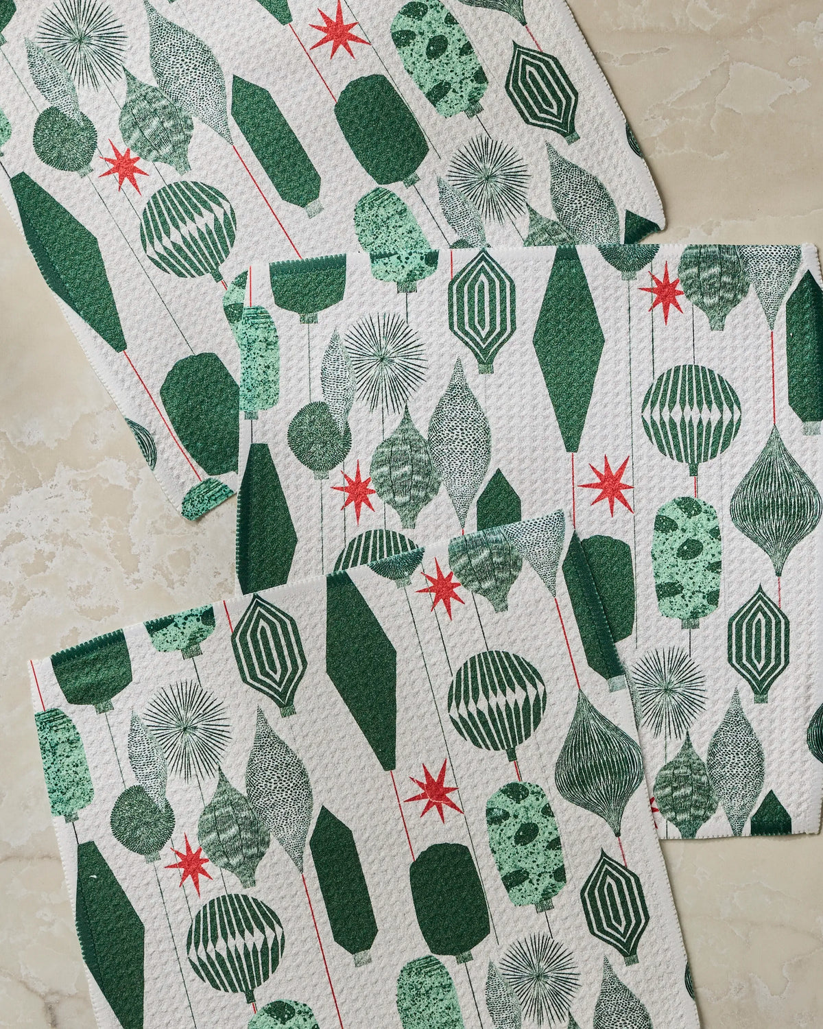 Four white recycled dishcloths from the Geometry Alpine Revelry set, featuring green geometric and leaf patterns with red starbursts, are arranged overlapping each other on a light beige marble surface, creating a charming and eco-friendly aesthetic.