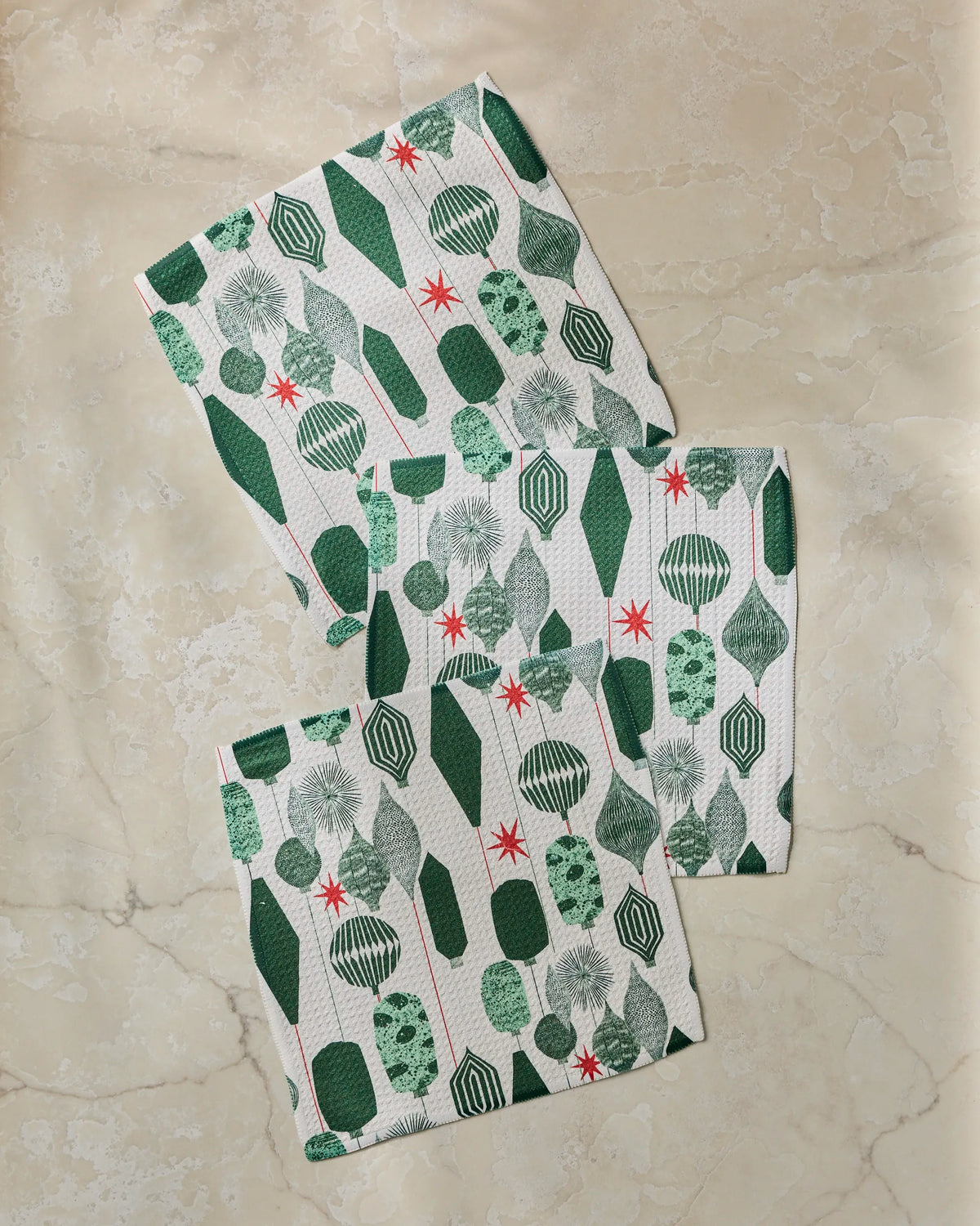 Three Alpine Revelry paper napkins from Geometry, featuring green and white geometric and starburst patterns reminiscent of a stylish dishcloth set, are arranged on a beige marble surface.