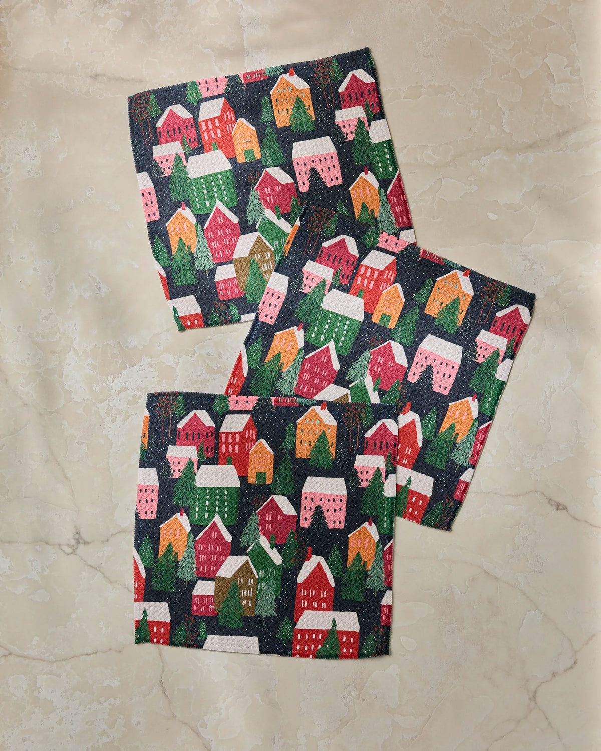 Three festive napkins from Geometry’s Alpine Village collection, featuring a vibrant pattern of red, pink, green, and yellow houses and trees arranged on a marble surface. Made from recycled materials, this cheerful set of super-absorbent dishtowels brings the charm of a cozy holiday village to your celebrations—combining style with sustainability.