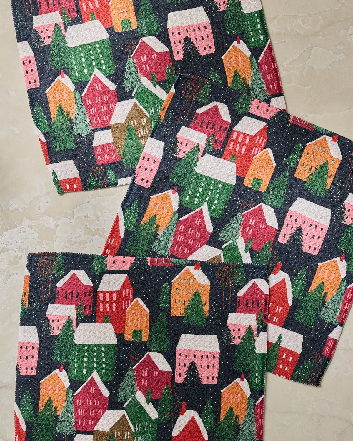 Three Alpine Village napkins from Geometry, crafted from recycled materials, feature a charming pattern of red, pink, and green houses and trees dusted with snow, all laid on a beige marble surface.