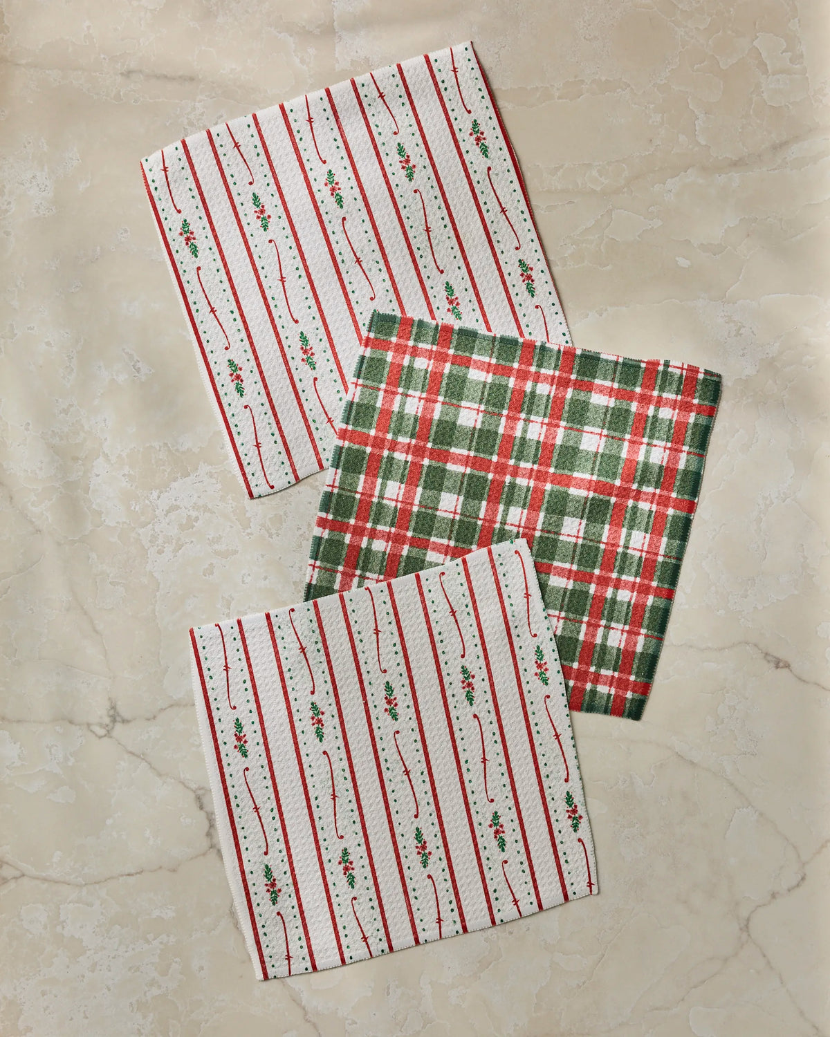 Three festive Geometry “Alpine Winters” cloth napkins are displayed on a light marble surface: two showcase red and green holly stripes on white, while the third features a classic red, green, and white plaid pattern—perfect for bringing holiday cheer to your dishcloth set or pairing with other absorbent dishcloths in your kitchen.