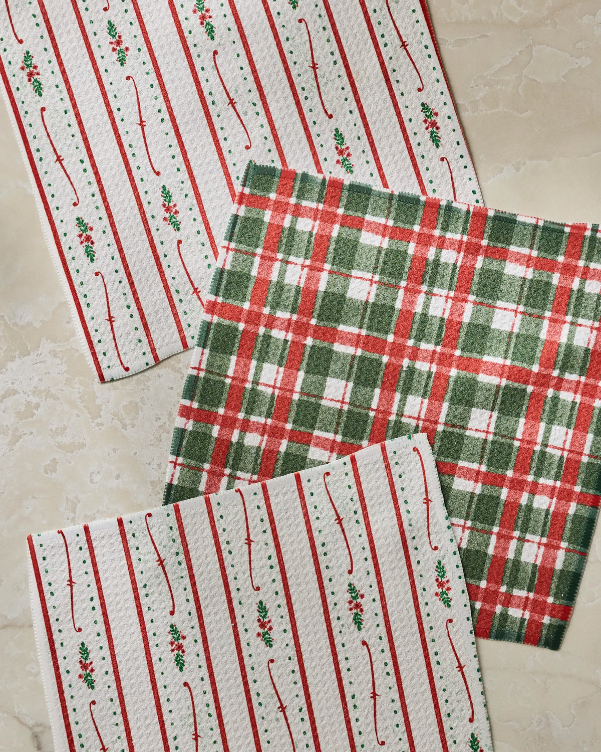 The Geometry Alpine Winters dishcloth set features three absorbent cloths—two with red and green holly and stripes on white backgrounds, and one in a red, green, and white plaid pattern—all arranged on a beige surface to bring holiday cheer and convenience to your kitchen.