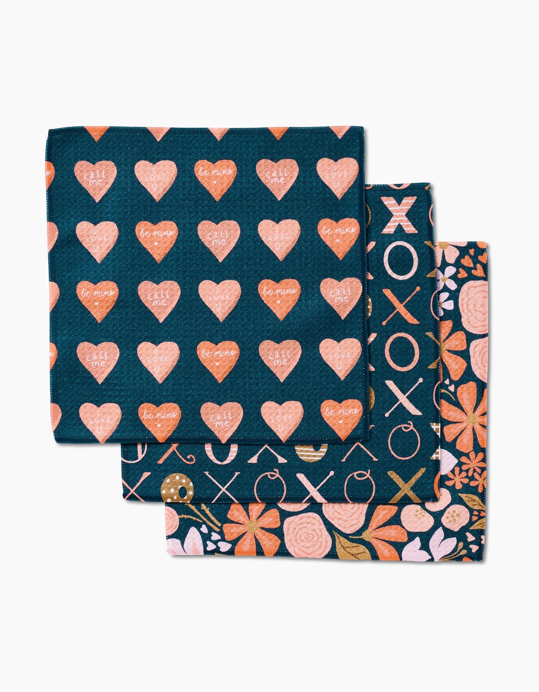 Amandine by Amy MacCready: Set of three Geoweave fabric napkins with Valentines designs—coral hearts and phrases, XOXO circles, and heart-shaped flowers—on a dark blue background. Stylish, eco-friendly dishcloths.