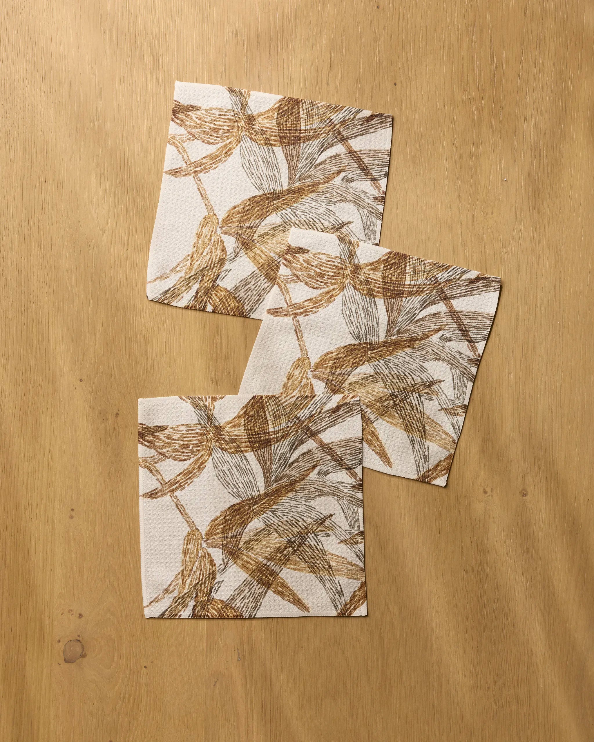 Three Amber Wildflower napkins by Geometry, featuring a brown and beige leafy pattern, are arranged diagonally on a light wooden surface with visible wood grain. The napkins overlap slightly, evoking the charm of recycled dishcloths.