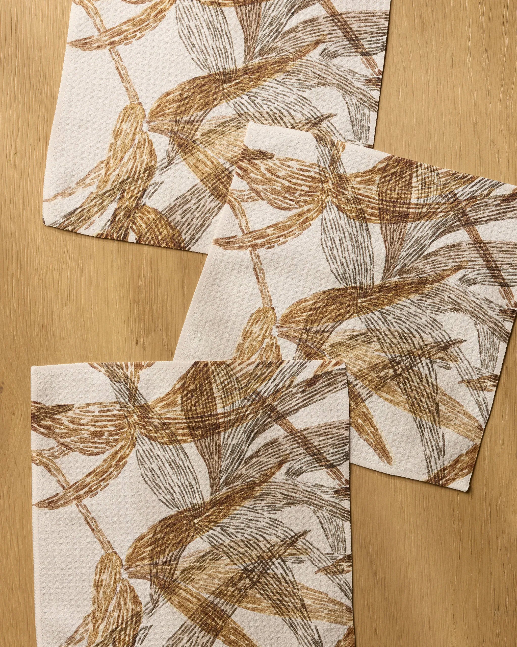 Three Amber Wildflower napkins from Geometry, featuring a brown and beige abstract leaf design, are displayed on a light wood surface—perfectly complementing any recycled dishcloths or absorbent dish towels in your kitchen.