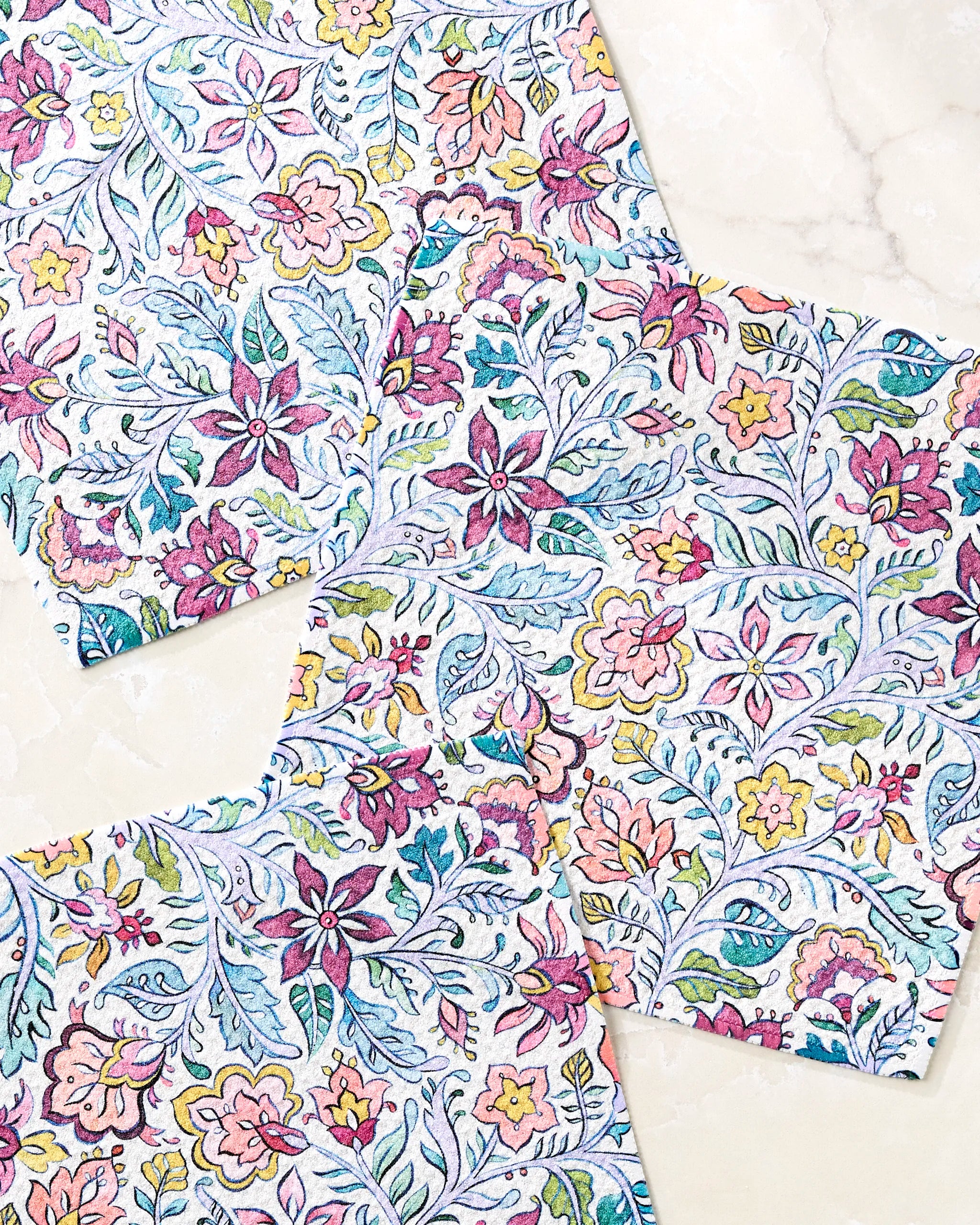 Three Amethyst napkins by Maayan Alper-Swan, made from recycled materials, display intricate pink, yellow, blue, and green botanical patterns on a light marble surface. The square napkins overlap slightly, highlighting their vibrant designs.