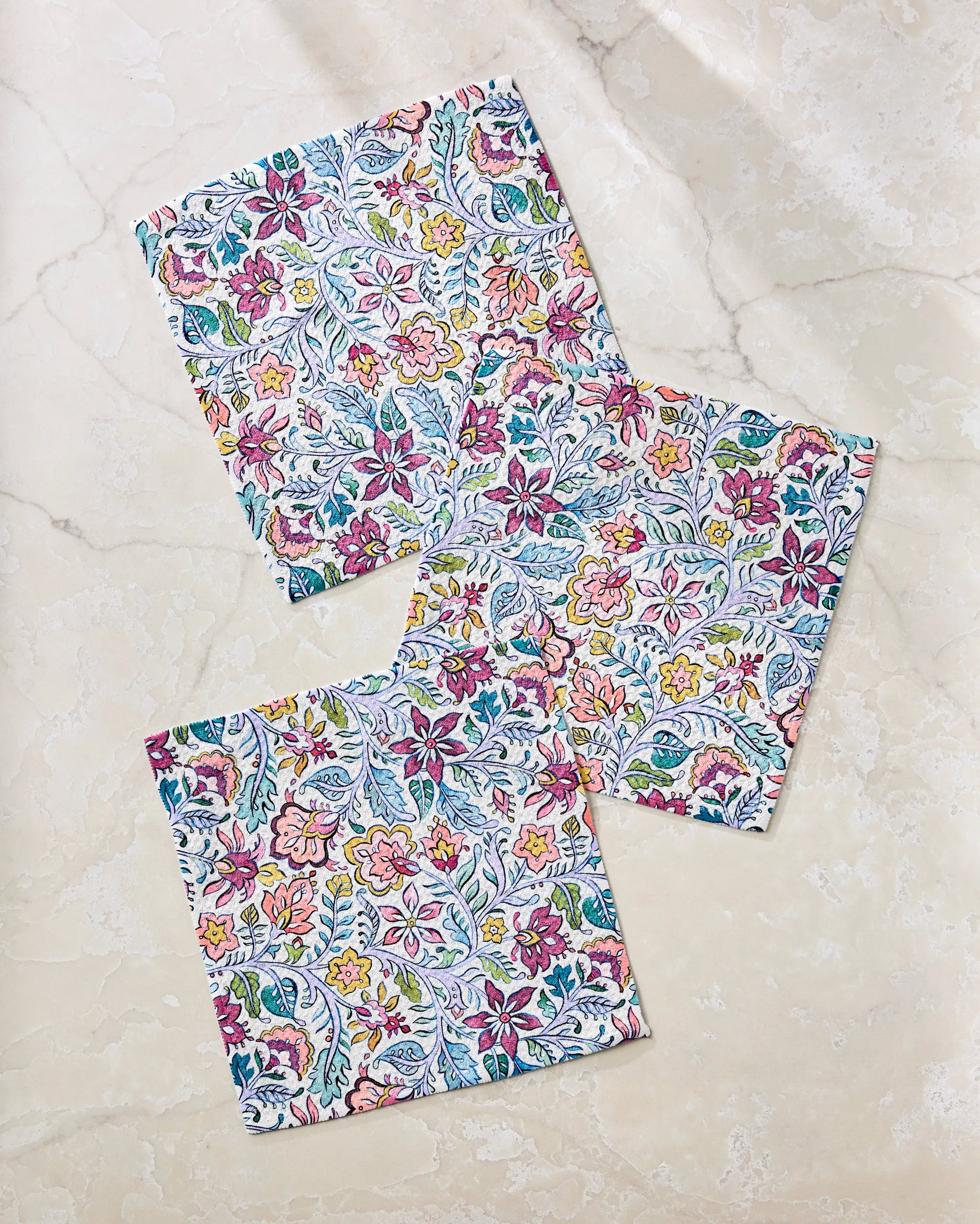 Three “Amethyst” square napkins by Maayan Alper-Swan, made from recycled materials, feature a vibrant floral and leafy design with pink, yellow, blue, and green accents. They’re arranged on a light marble surface.
