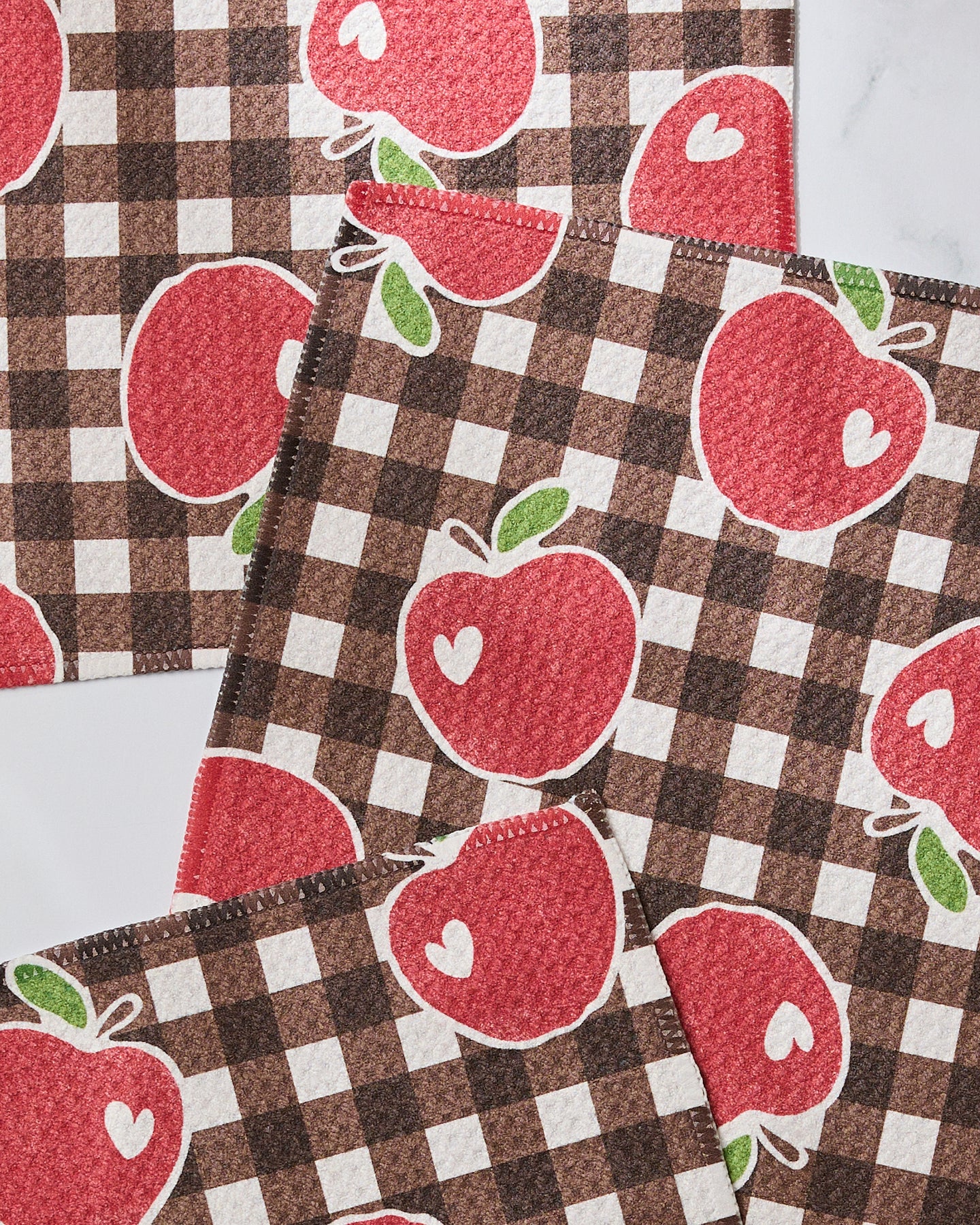 Three brown and white checkered dishtowels from Geometrys “Apple Picking Afternoon” set feature large red apples with small white hearts. The overlapping set is displayed on a white surface.