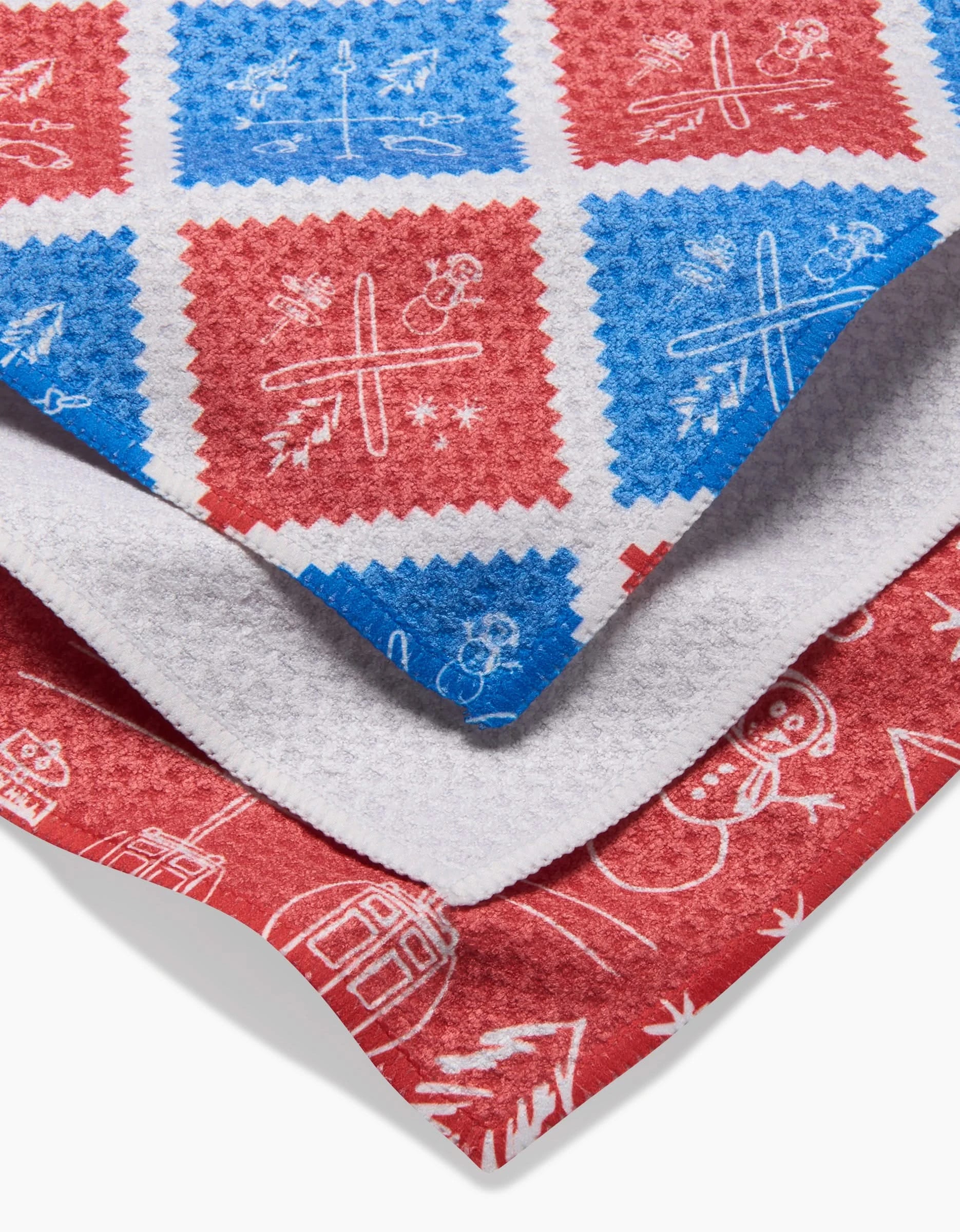 Three festive Apres Ski kitchen towels by Rosa Kichline, stacked together, feature red, white, and blue holiday designs with snowmen, Christmas trees, and snowflakes on soft-textured Geoweave fabric.