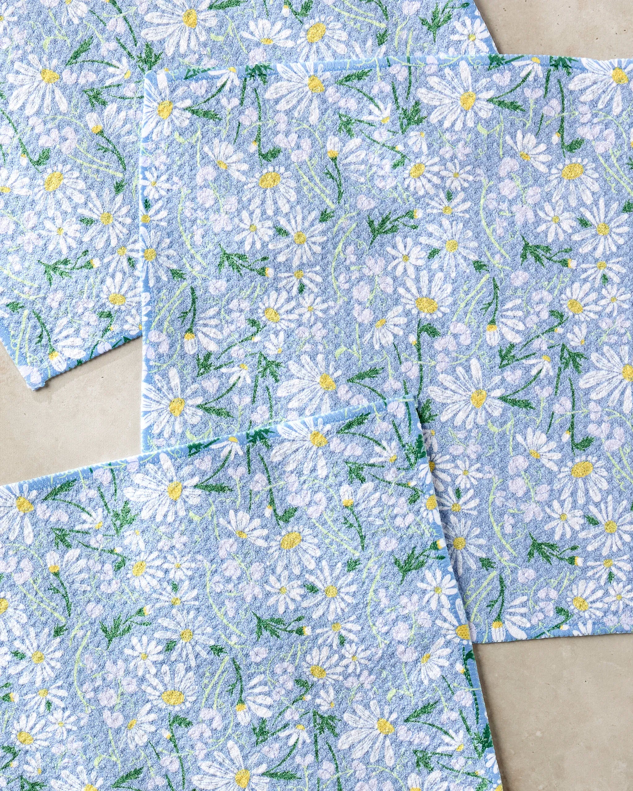 The April napkins by Elisabeth Wing are a set of four blue, recycled-material napkins featuring white daisies with yellow centers and green stems, arranged in an overlapping design on a neutral background.