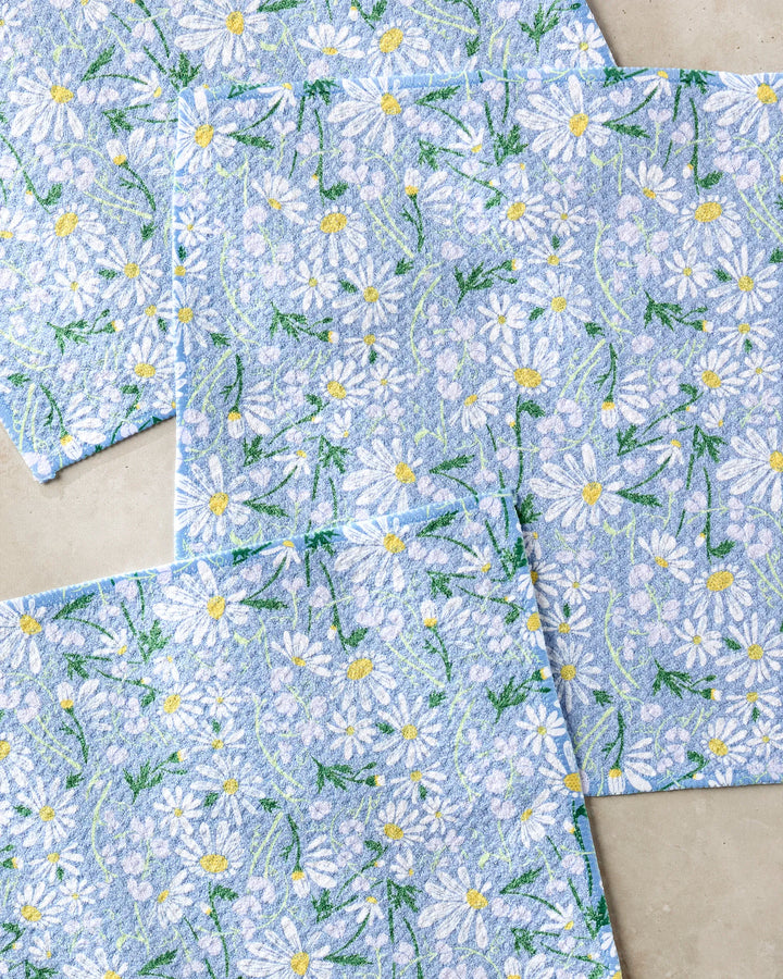 The April napkins by Elisabeth Wing are a set of four blue, recycled-material napkins featuring white daisies with yellow centers and green stems, arranged in an overlapping design on a neutral background.