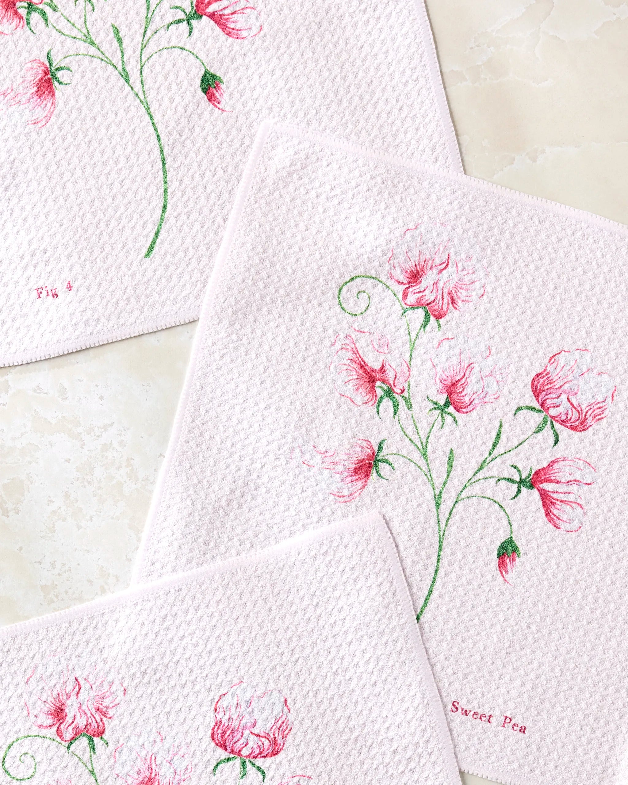 The Linda Fox "April Sweet Pea" set includes three pale pink recycled dishcloths with pink and green floral embroidery—two feature sweet pea flowers labeled Fig 5 and Sweet Pea—offering a charming, absorbent touch to your kitchen.
