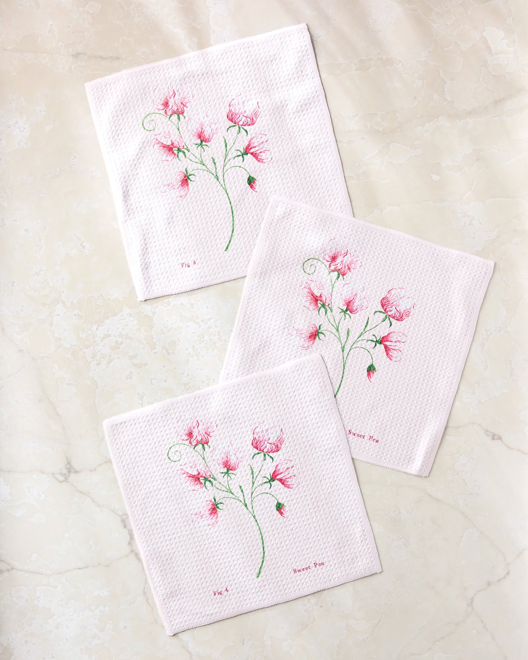 Three white textured napkins from the Linda Fox April Sweet Pea set display delicate pink flower embroidery with handwritten text and green stems, arranged on a marble surface. Each napkin features a similar floral design.