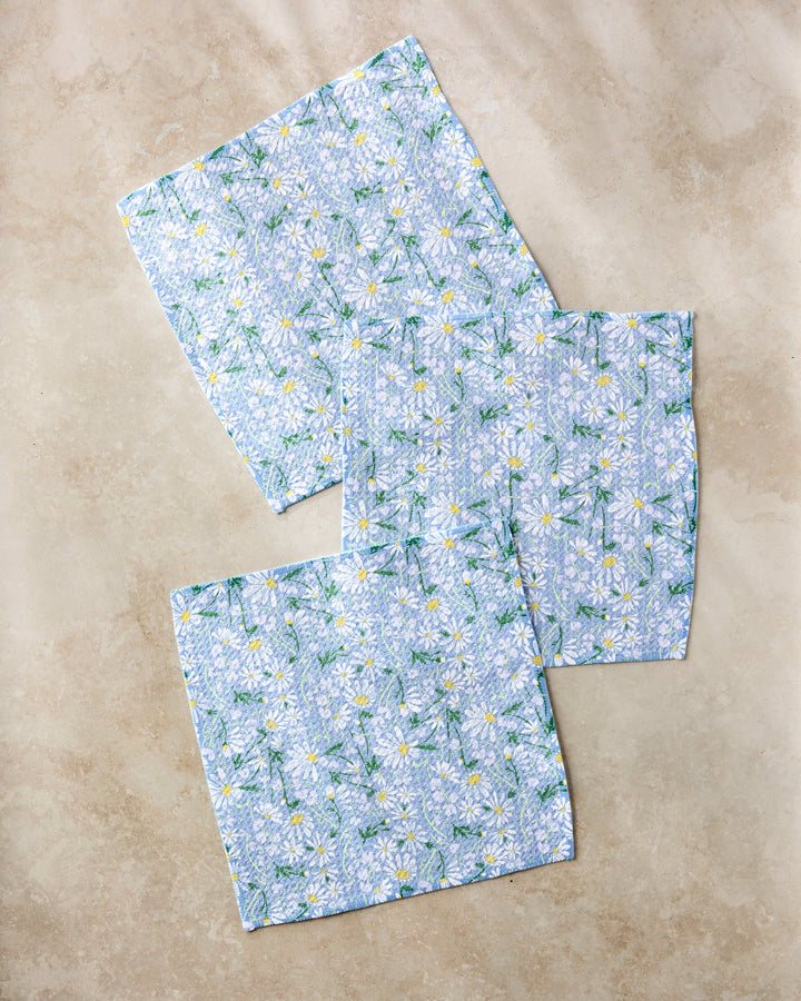 Three Elisabeth Wing "April" napkins, made from recycled materials and featuring a blue background with white daisy flowers, are arranged on a beige surface.