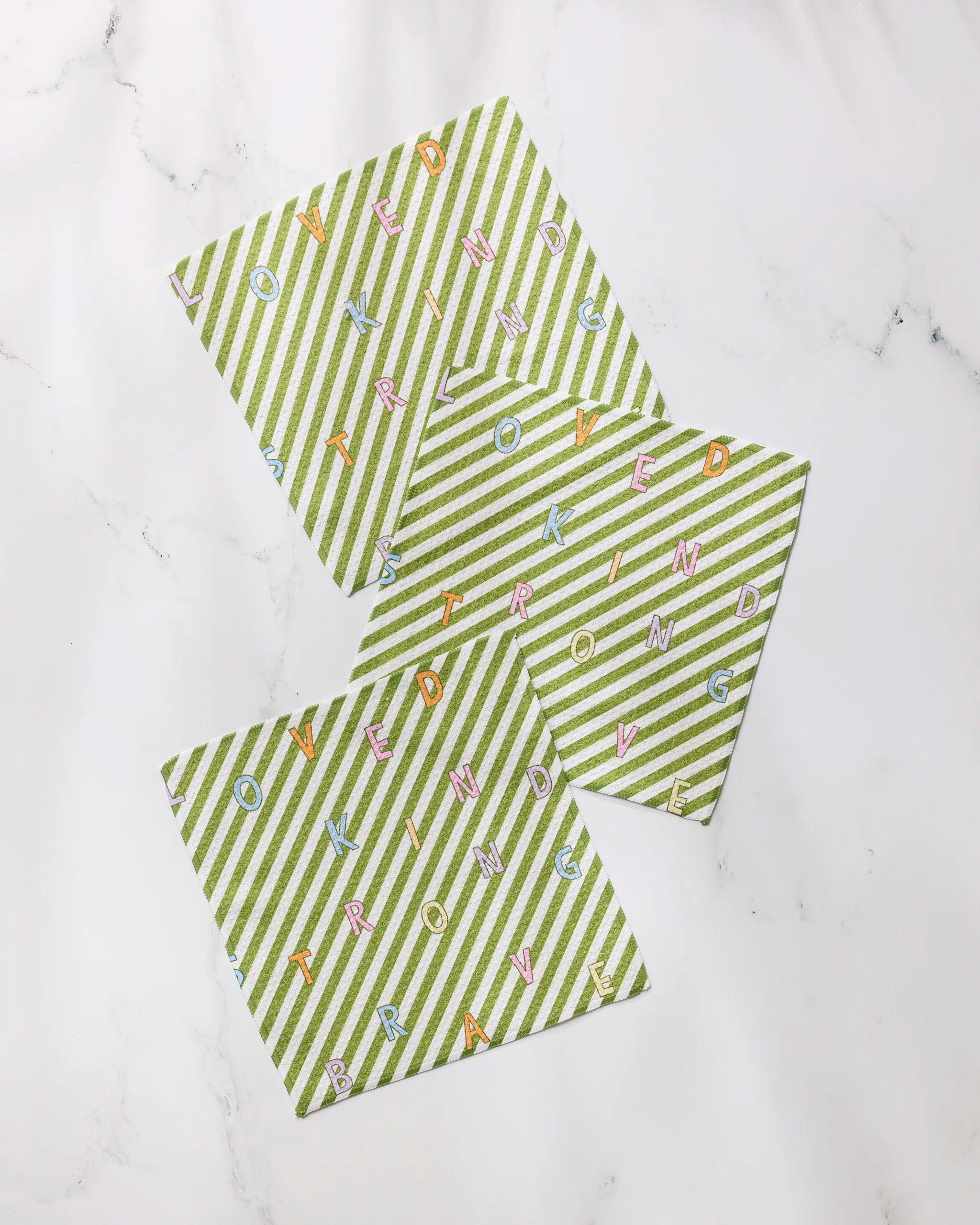 Three green and white striped Geometry Attributes cards are arranged on a white surface, with colorful letters scattered across them forming the words KIND, LOVED, BRAVE, and STRONG in various directions.