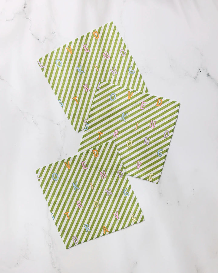 Three green and white striped Geometry Attributes cards are arranged on a white surface, with colorful letters scattered across them forming the words KIND, LOVED, BRAVE, and STRONG in various directions.