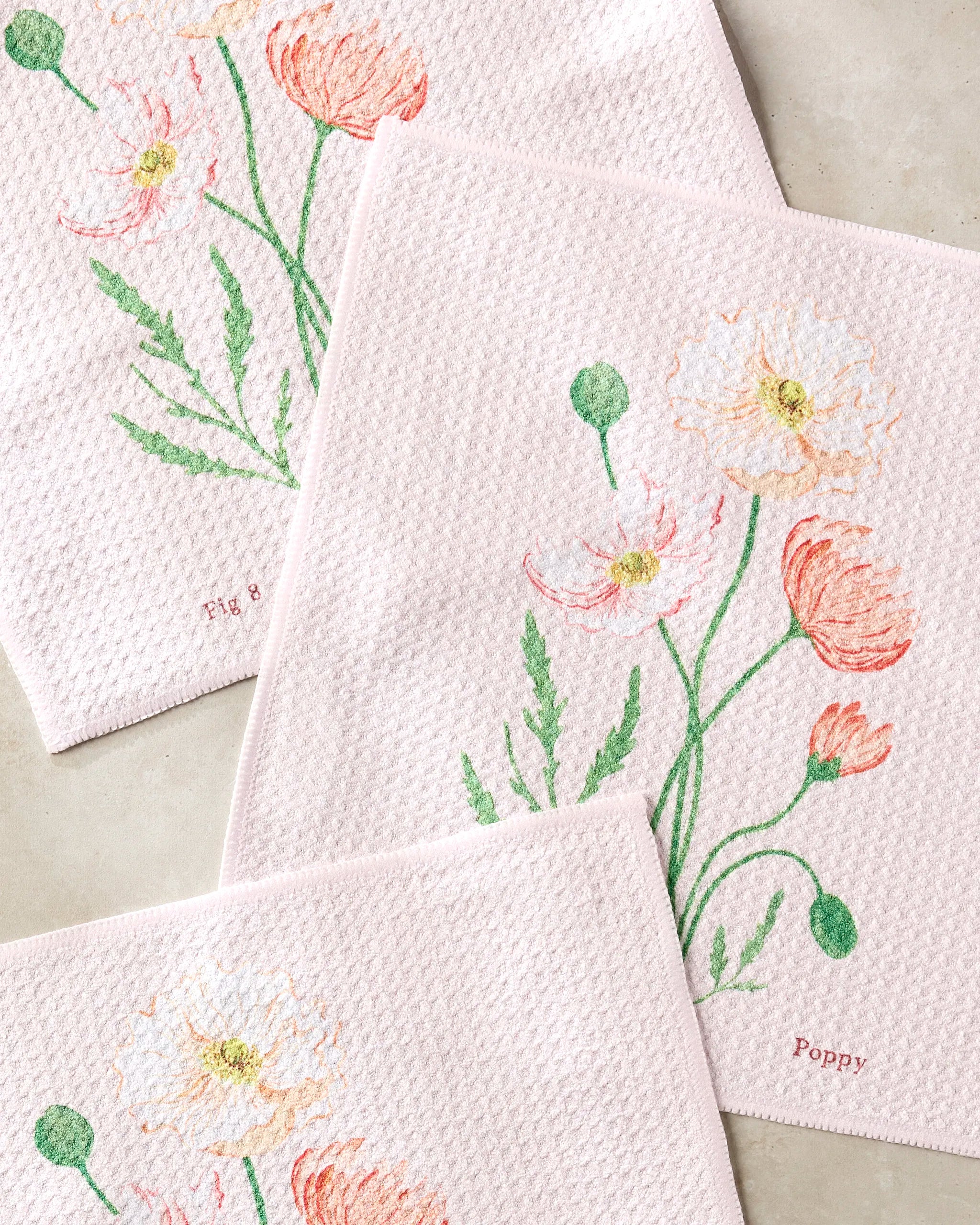 The Linda Fox August Poppy set includes three pale pink textured cloths with colorful poppy illustrations and green stems, "Poppy" text on one, and is crafted from recycled materials for super absorbency.