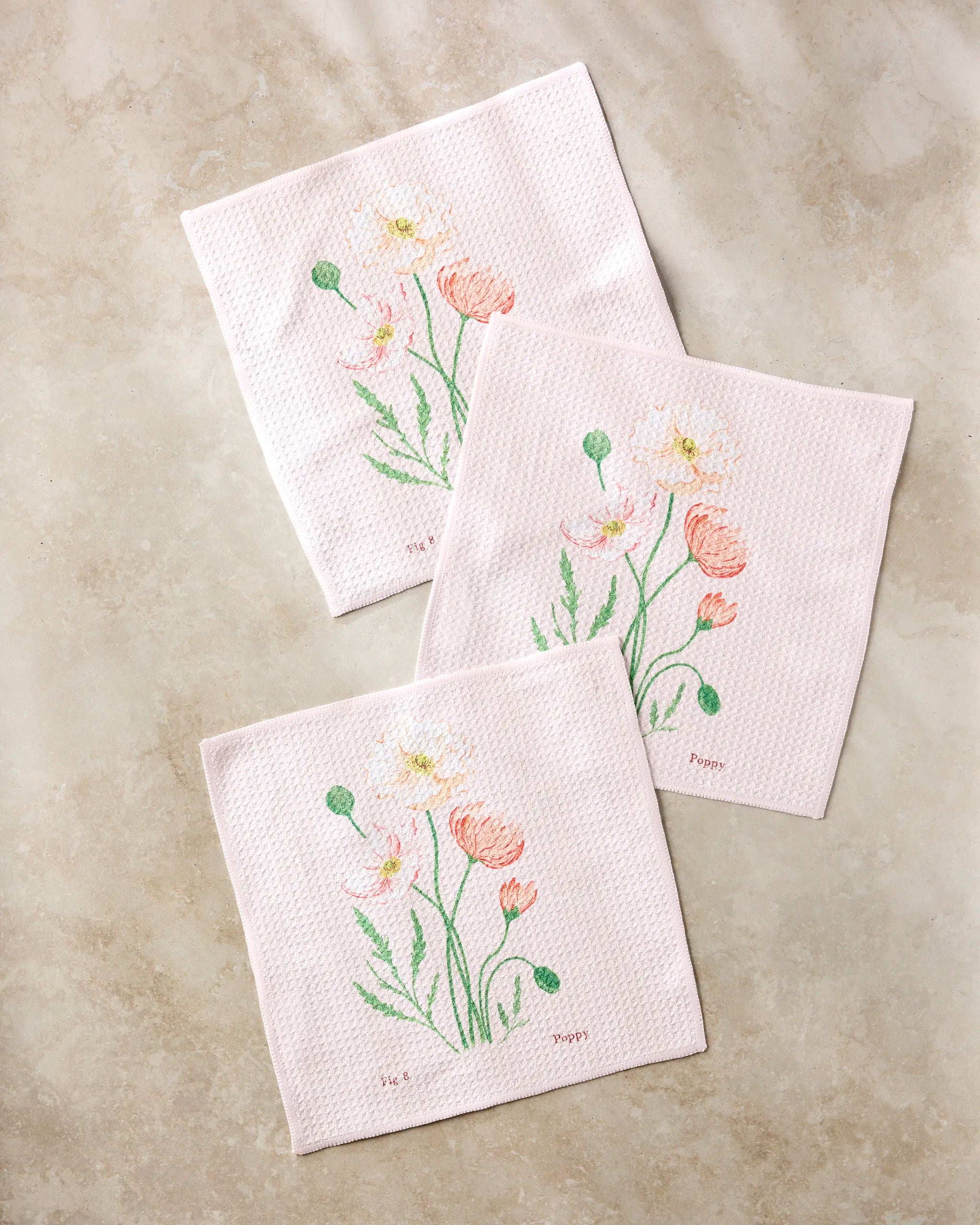 Three August Poppy cloth napkins by Linda Fox, made from recycled materials, feature pale pink fabric with embroidered pink and white flowers and green stems, shown arranged on a light beige surface.