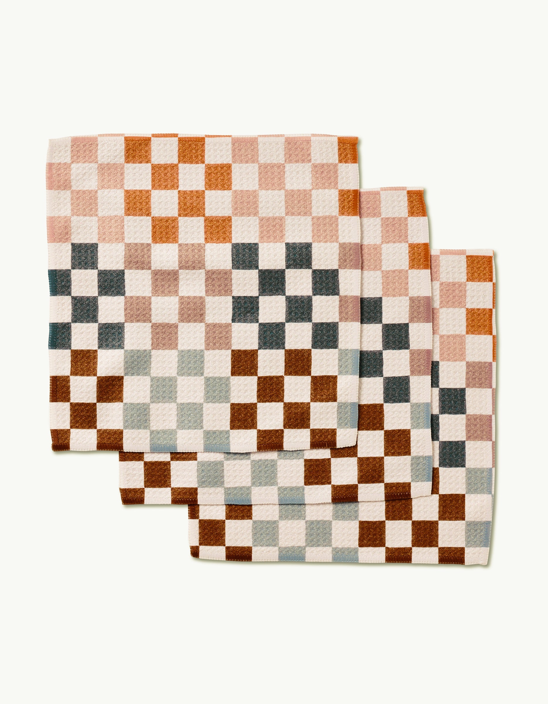 Three GEOMETRY Autumn Checkers napkins, made of Geoweave fabric with a brown, orange, pink, cream, and gray checkered pattern, are neatly stacked on a light background.