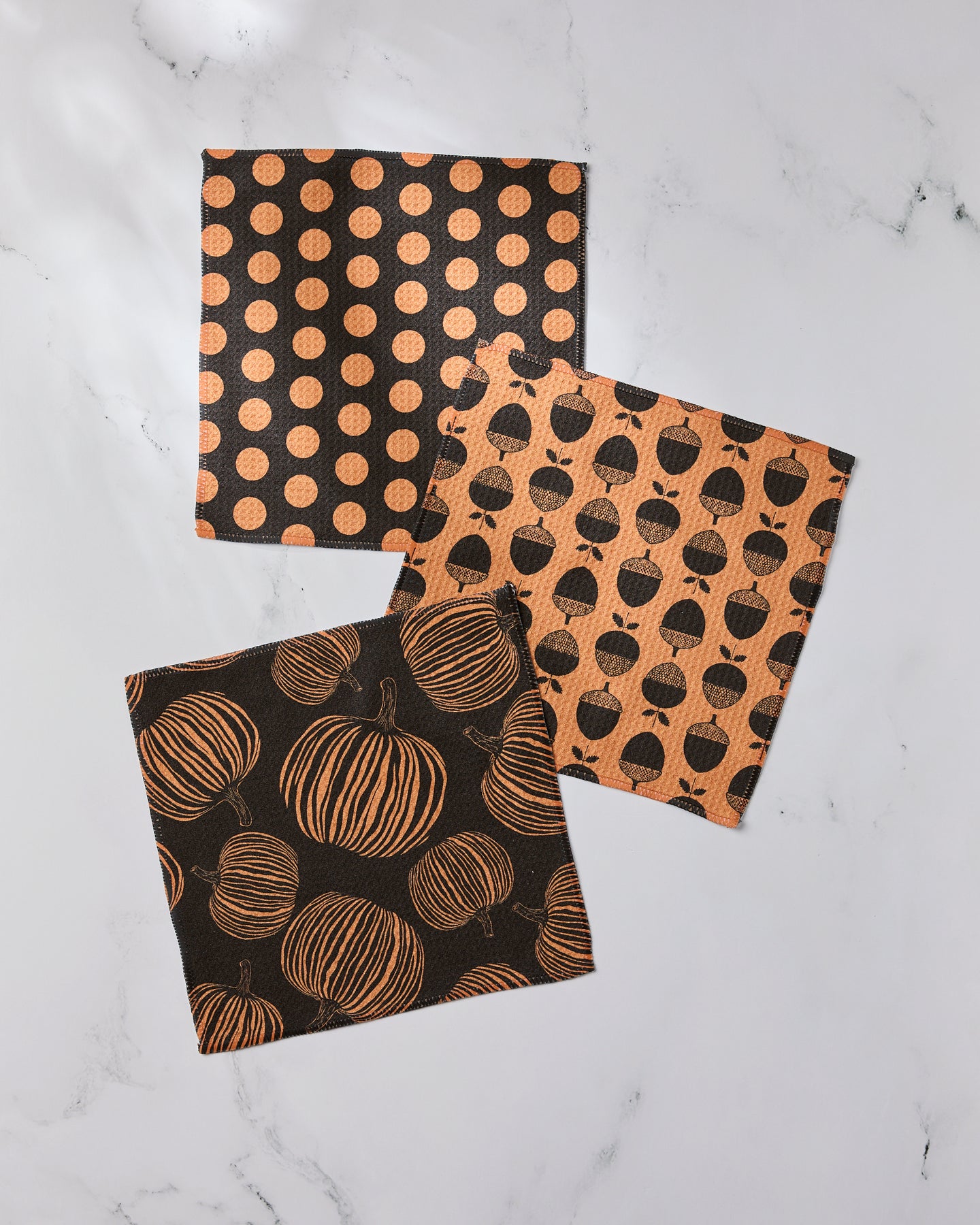 Autumn Noir by Geometry: Three square napkins made from recycled materials feature Halloween patterns—orange polka dots on black, black pumpkins on orange, and orange pumpkins on black—displayed on white marble.