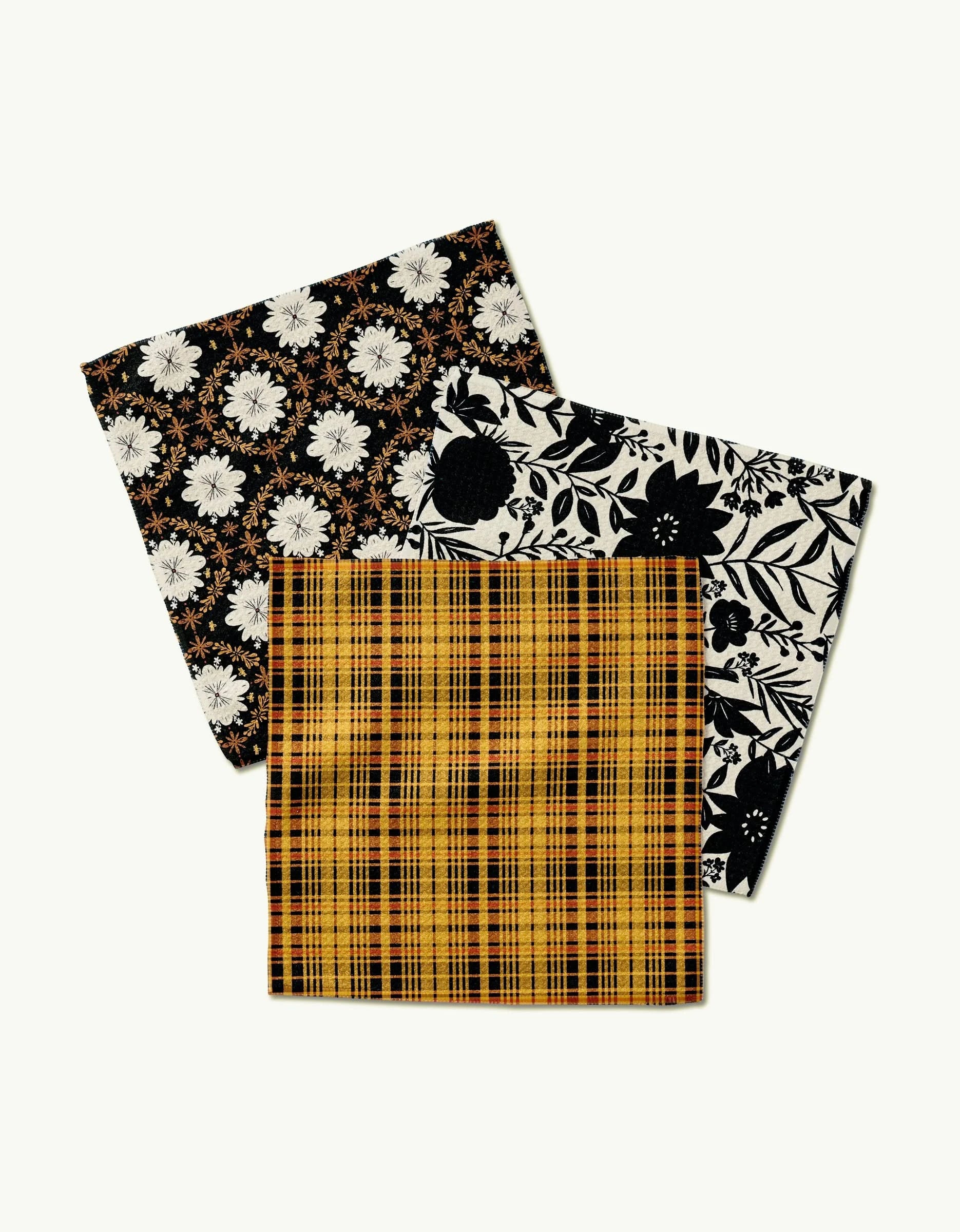 Three folded Danelys Sidron cloth napkins—Autumn Plaid in yellow and black plaid, plus two floral designs—are arranged overlapping on a light background. Made from Geoweave fabric for superior softness.