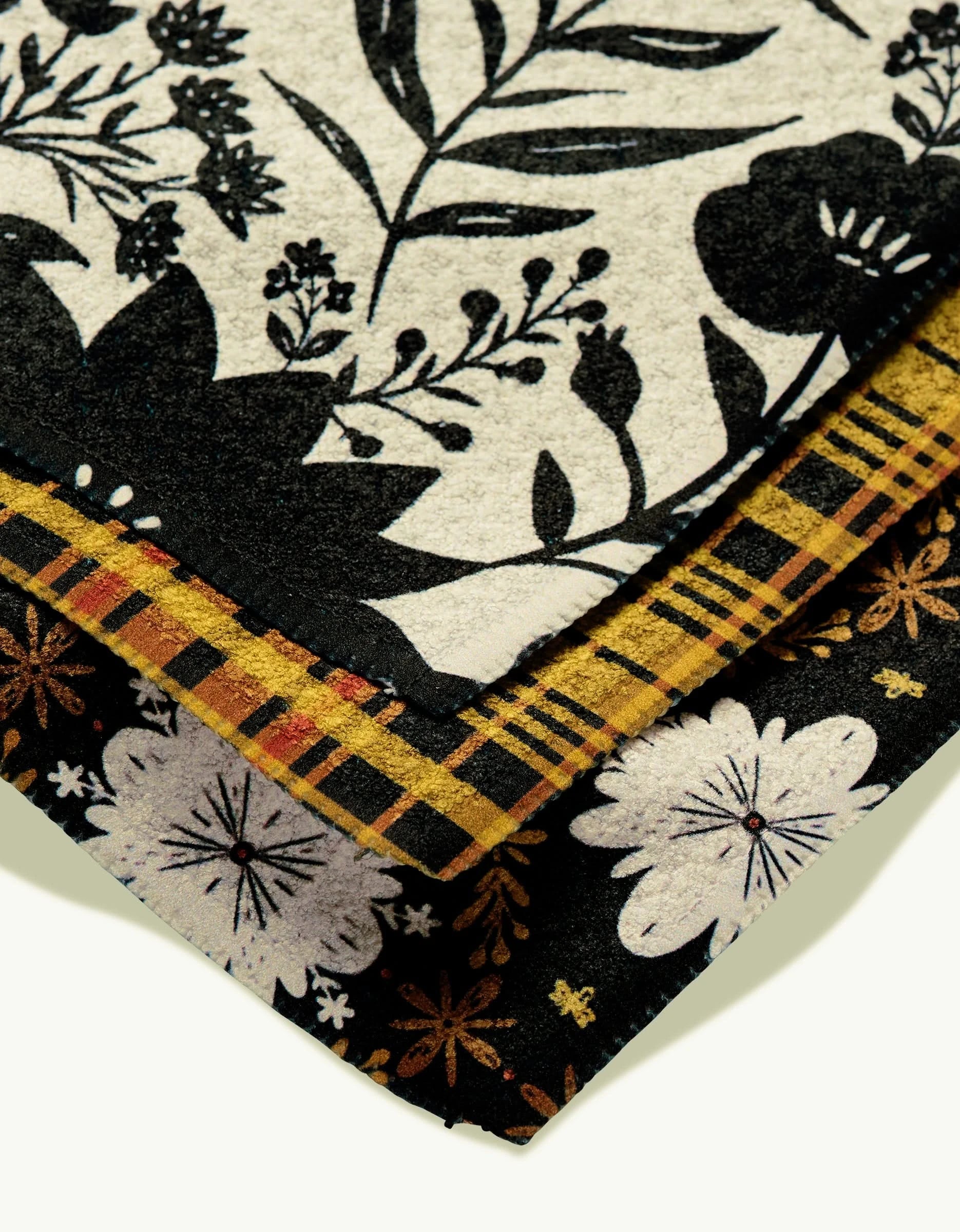 A close-up shows three overlapping rugs: the top has black florals on cream Geoweave, the middle is Danelys Sidrons Autumn Plaid with yellow and black plaid, and the bottom features white flowers on a black background.