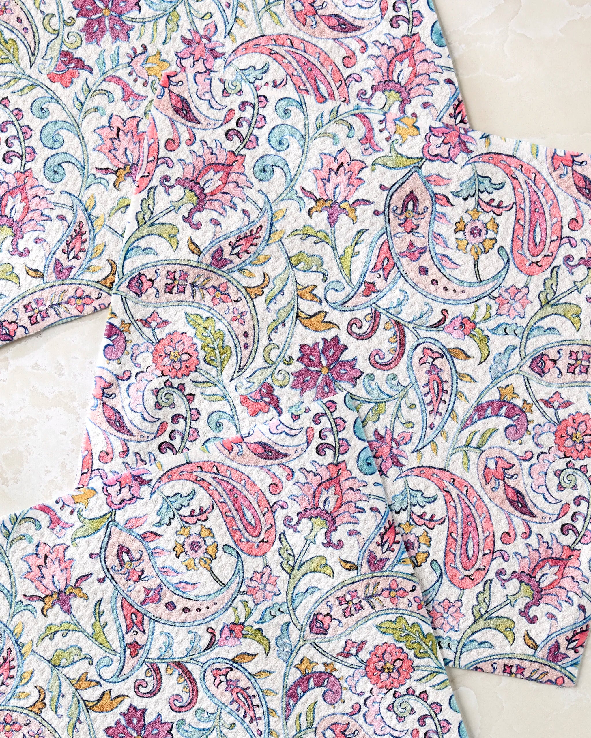 Three Bazaar dishcloths by Maayan Alper-Swan, featuring vintage-inspired floral and paisley patterns in pastel pink, blue, green, and purple, are shown overlapping on a light marble surface.