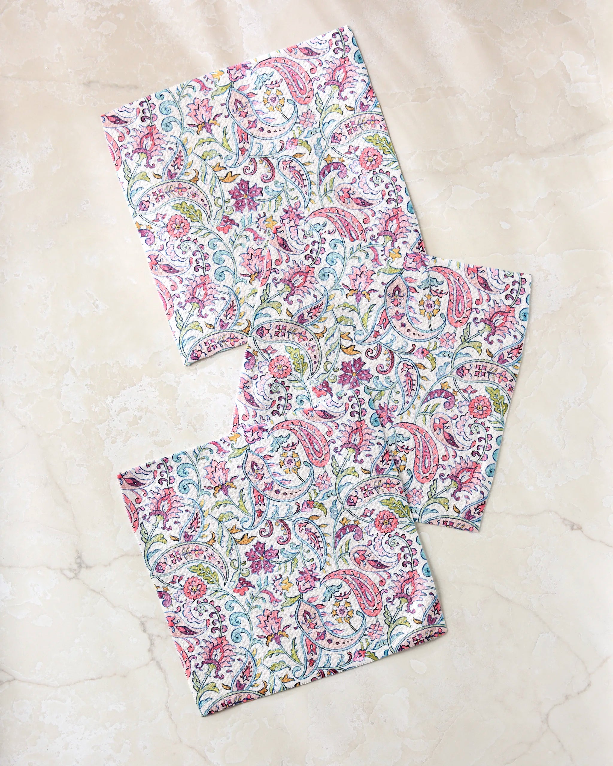Three pieces from the Bazaar dishcloth set by Maayan Alper-Swan, featuring colorful pink, green, blue, and yellow patterns, are arranged on a light beige marble surface.