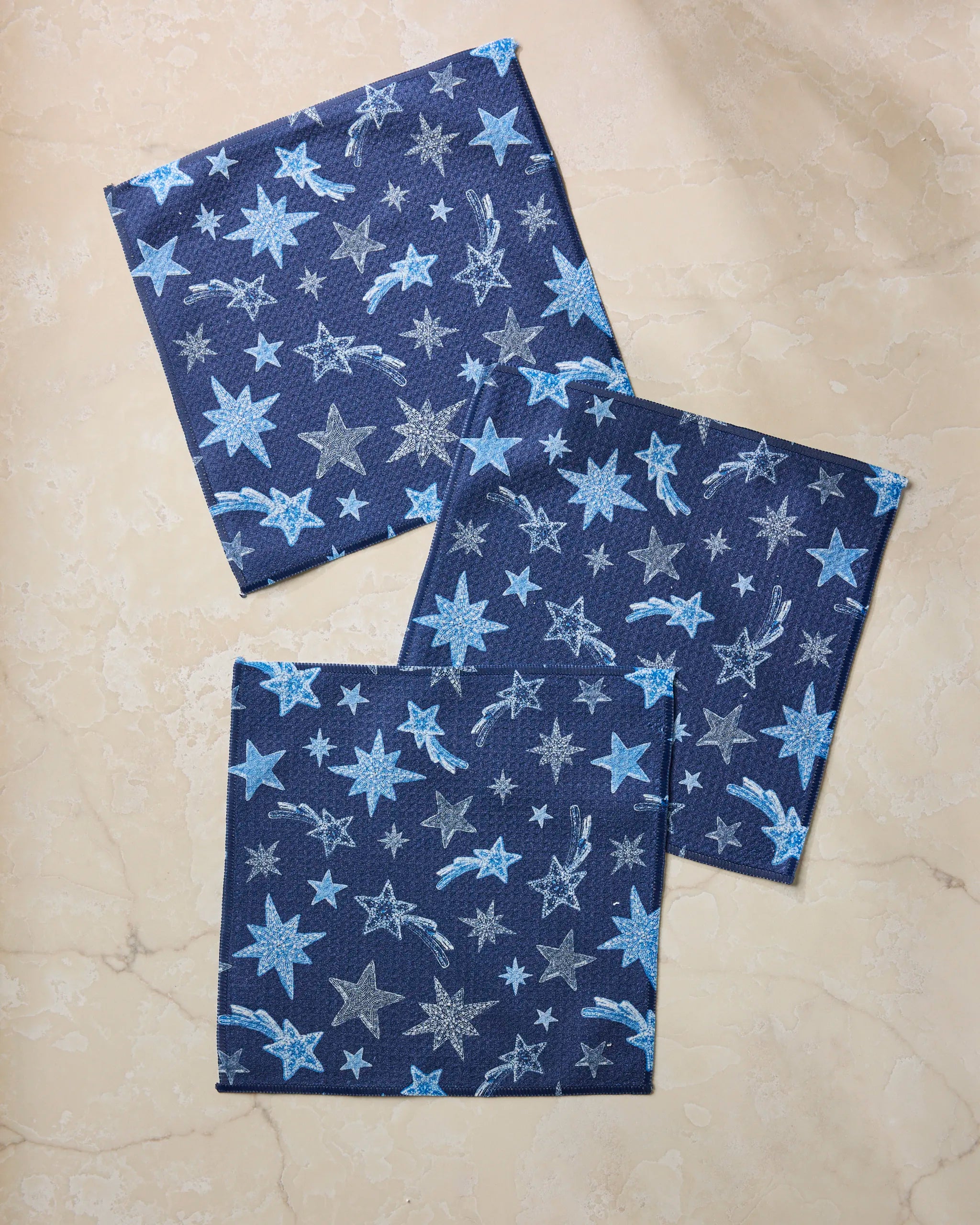 Three Geometry Beaded Stars Blue napkins, made from recycled materials, display light blue and white star patterns and are arranged diagonally on a light beige marble surface.