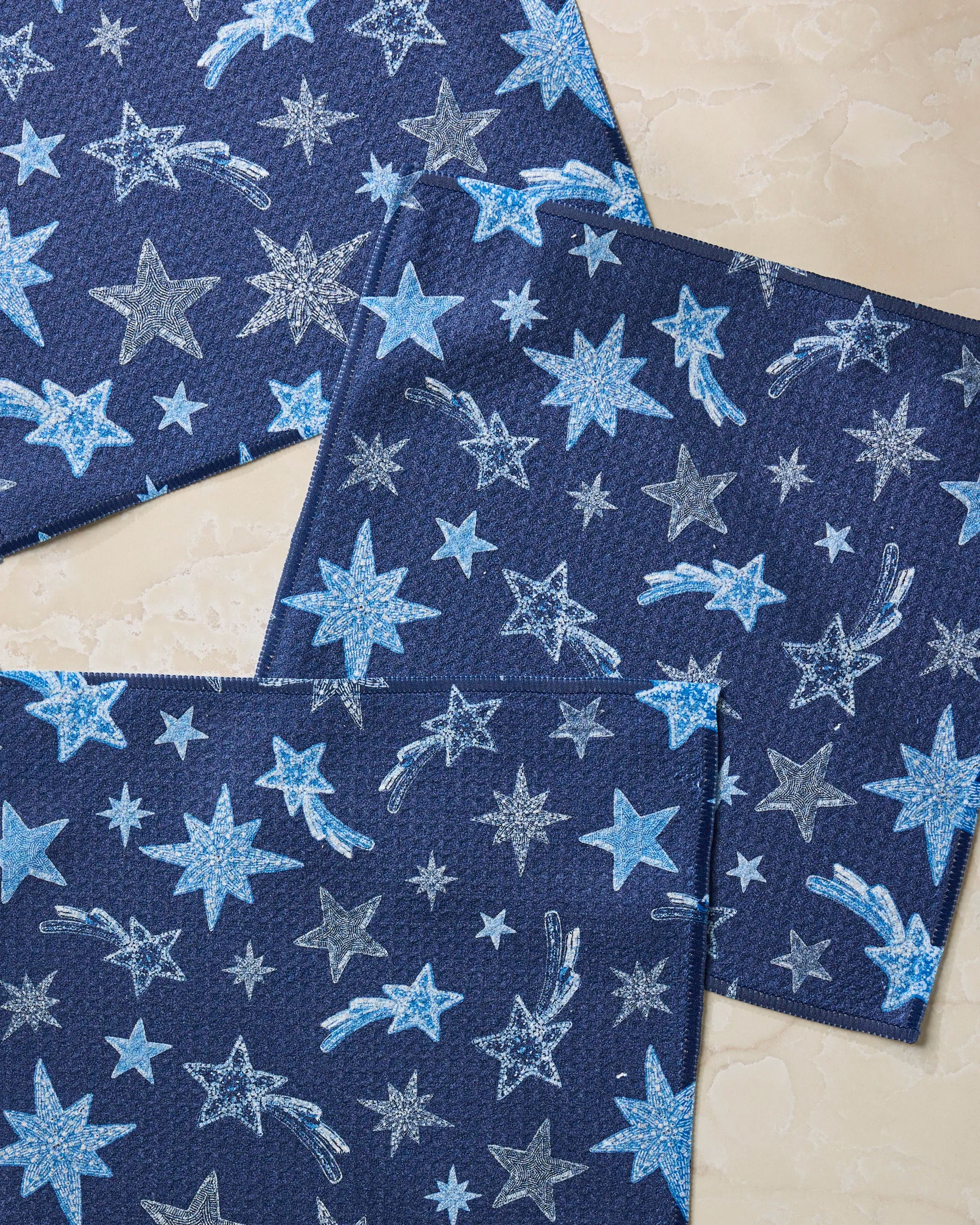 Geometry’s Beaded Stars Blue napkins, made from recycled materials and featuring sparkling blue and silver star patterns, are arranged on a light marble surface.