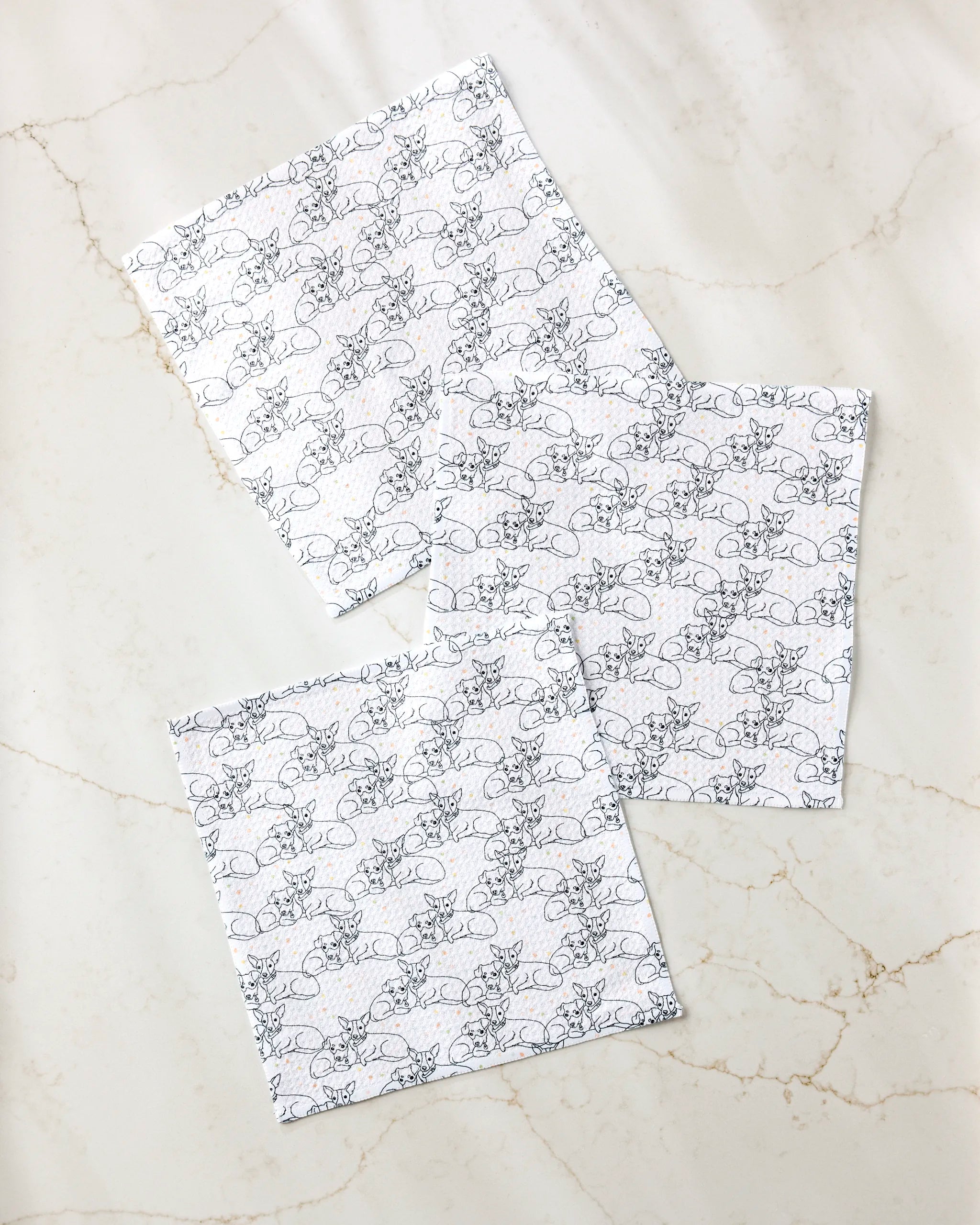 Three "Berry and Louie" napkins by Sweet Simple Vegan, featuring a black outlined floral print, are arranged on a light marble surface with beige veining. These are part of the brand's eco-friendly kitchen towel collection.