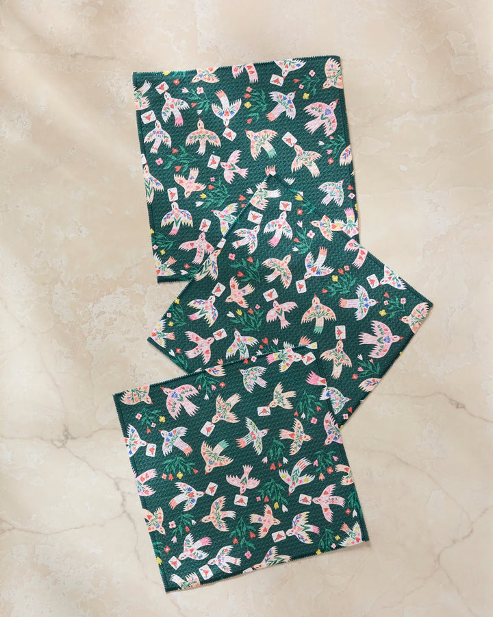 Three green "Birdsong Post" napkins by Ceyda Alasar, featuring a colorful bird and foliage design, are displayed on a light marble surface for a charming set.