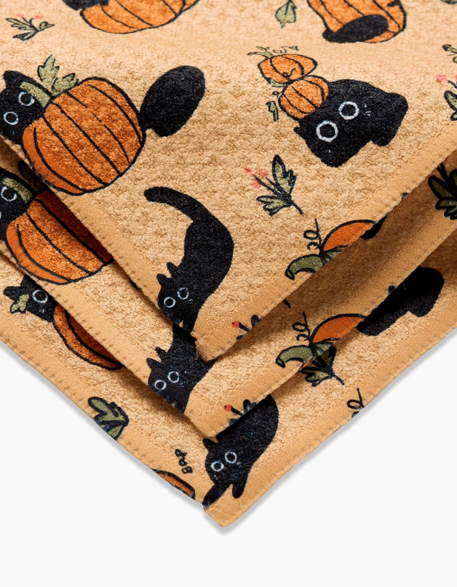 The Black Cats dishcloth by Janna Sue Design features a playful beige Geoweave fabric with black cats, orange pumpkins, and green leaves for a festive touch.