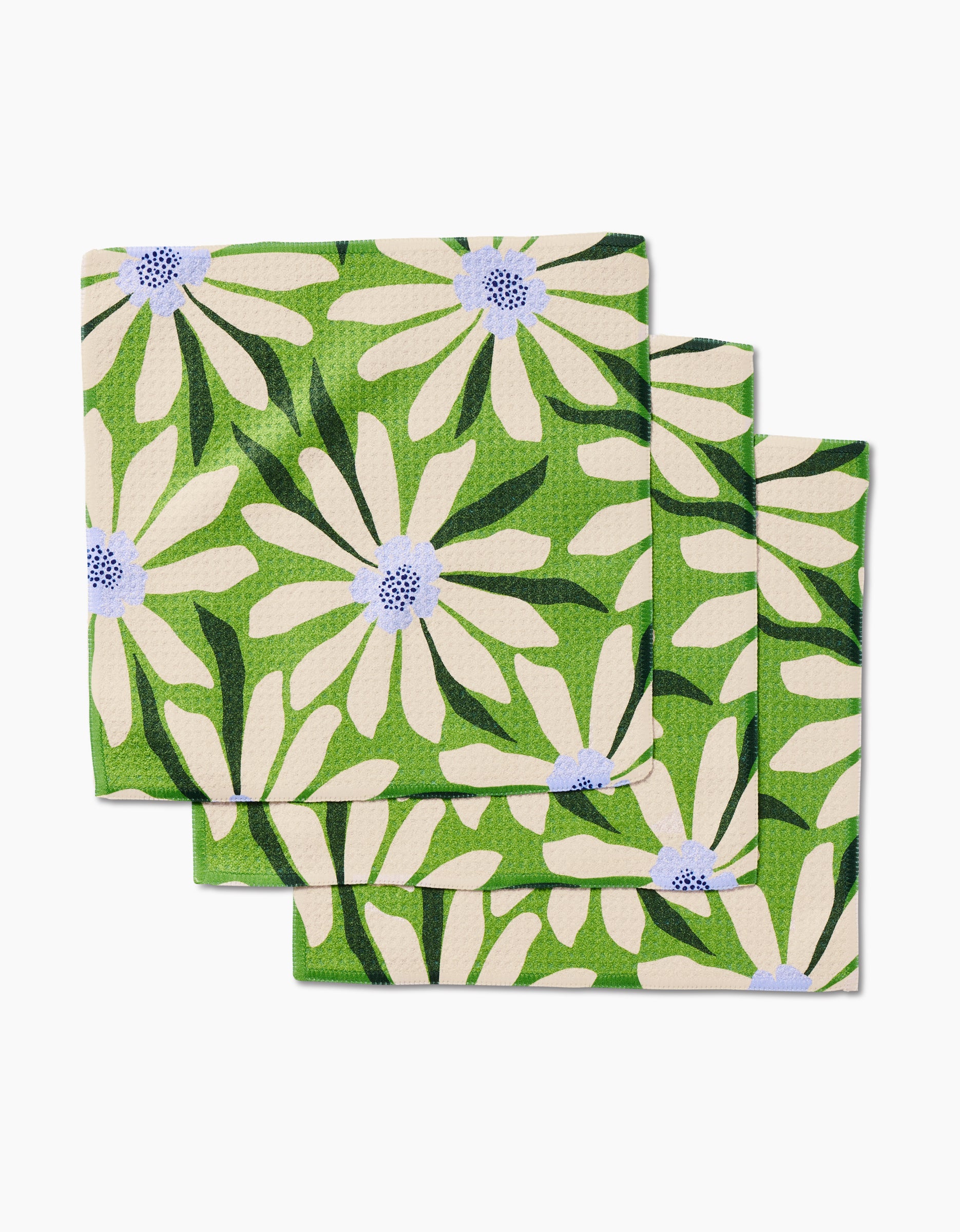 Three Bliss and Bloom napkins by GEOMETRY, made from recycled materials, feature large white daisies with blue and lavender centers plus dark green leaves on a green background, stacked in a slight overlap.