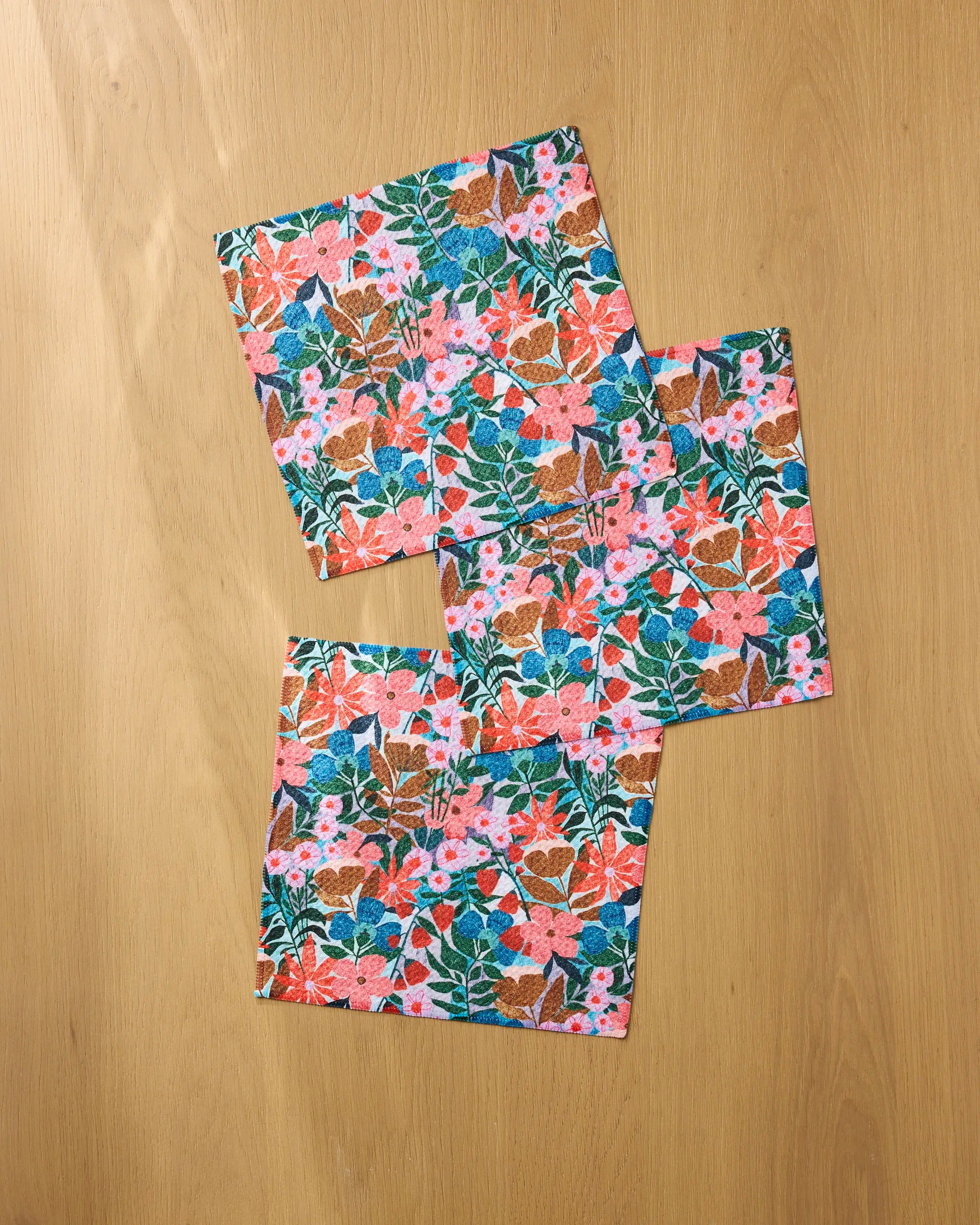 Three square Bloom Entangled napkins by Lisa Nelson, featuring a colorful floral pattern in pink, blue, orange, and green, are arranged overlapping on light wood—a charming accent for any dishcloth set.
