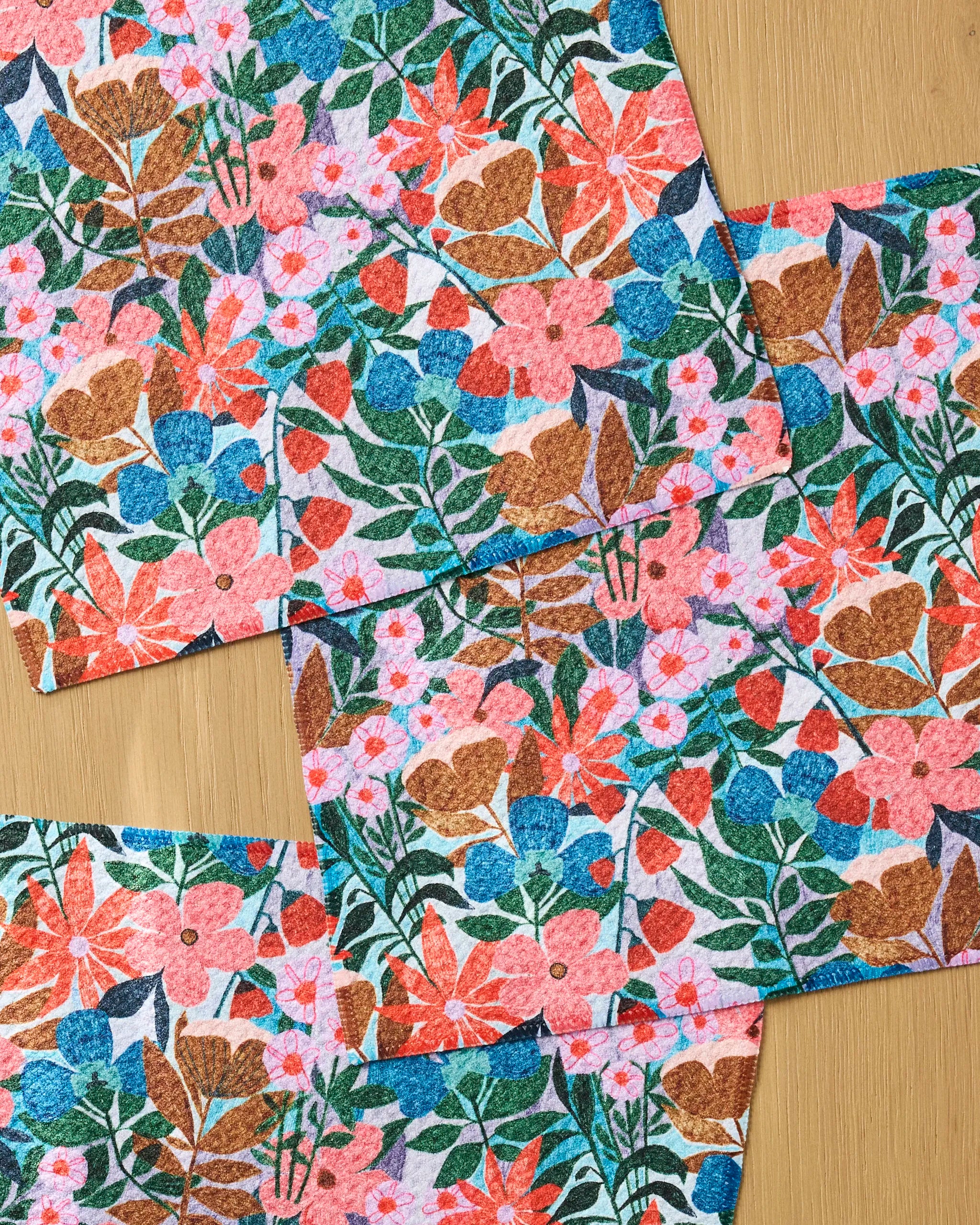 Four "Bloom Entangled" cloths by Lisa Nelson, made from recycled materials, feature vibrant pink, blue, red, and brown floral patterns with green leaves. They are arranged in overlapping squares on a wooden surface.