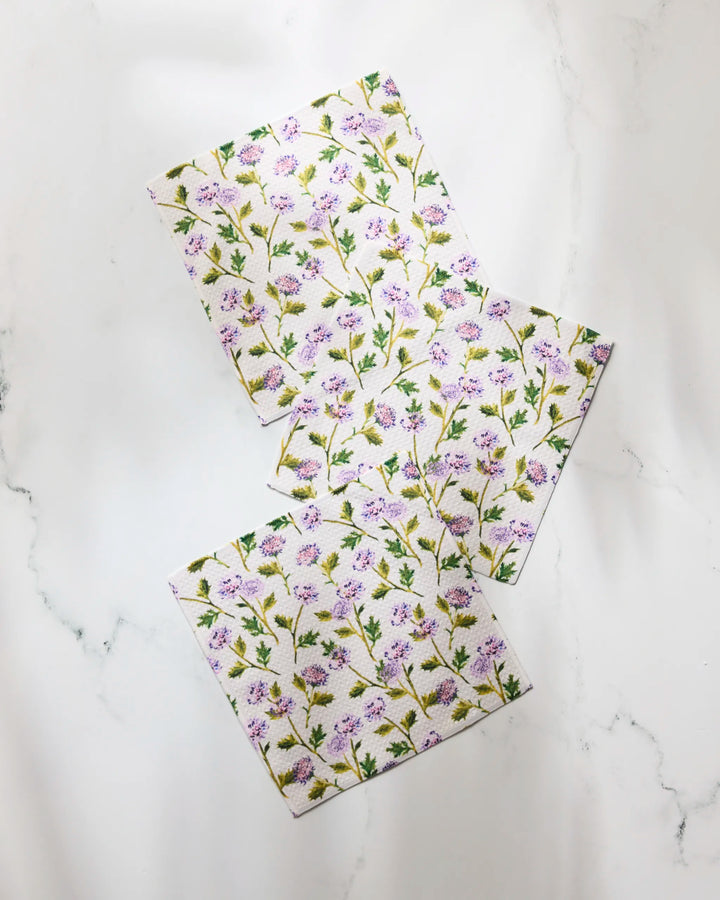Three Geometry Blooming Thistle cloth napkins, featuring green stems and tiny purple flowers, are arranged overlapping on a white marble surface.