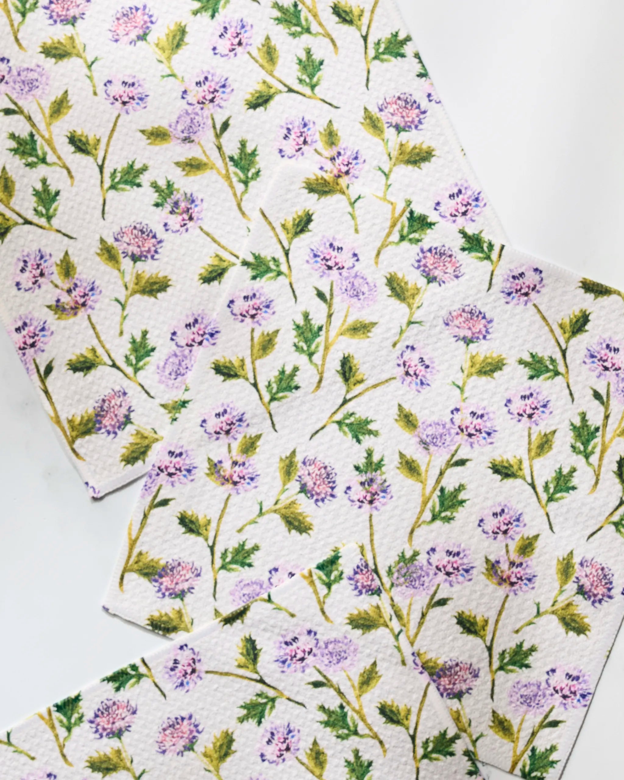 The Blooming Thistle dishtowel by Geometry features green stems, leaves, and small purple flowers on white. Super-absorbent and made from recycled materials for an eco-friendly touch.