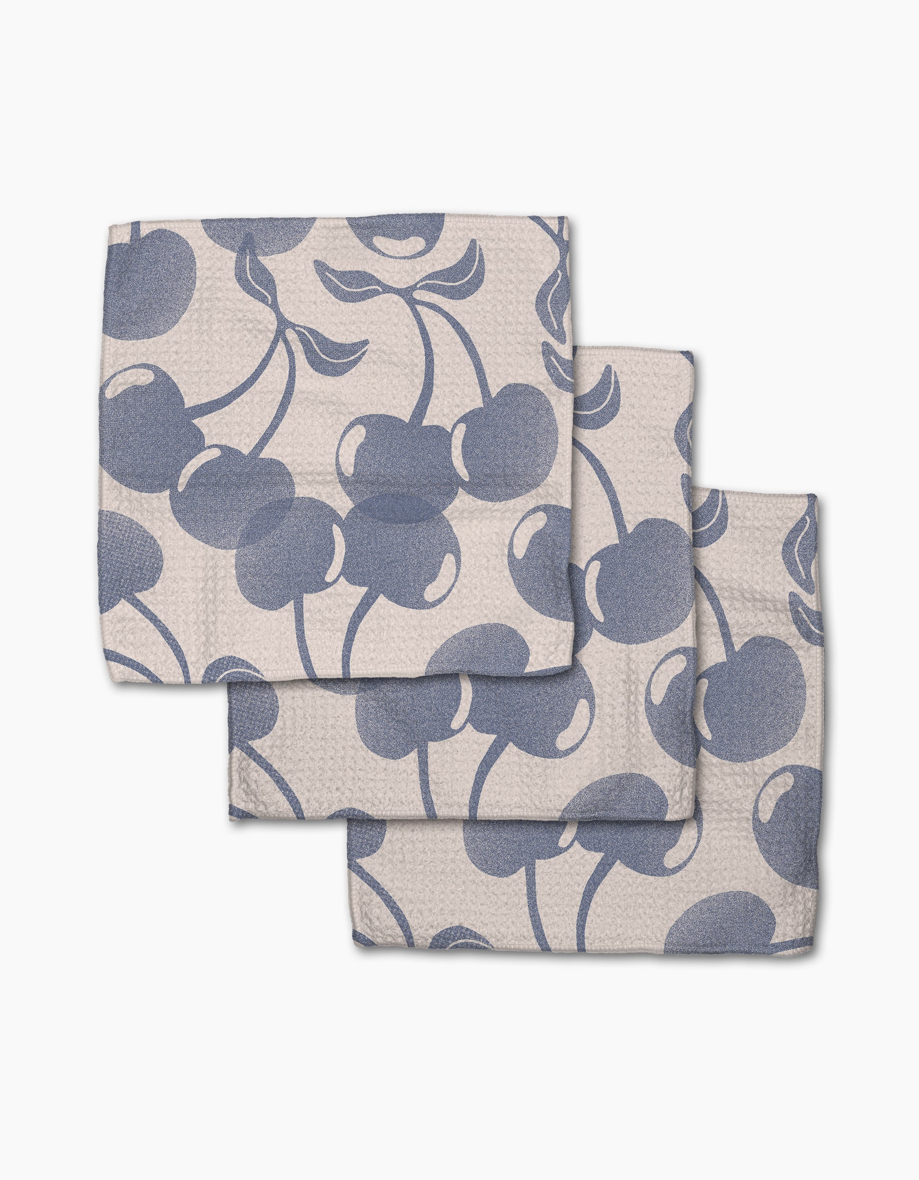 Three folded Geometry Blue Cherry Dreams beige cloth napkins, made from recycled materials with a blue cherry and leaf pattern, overlap each other on a white background.
