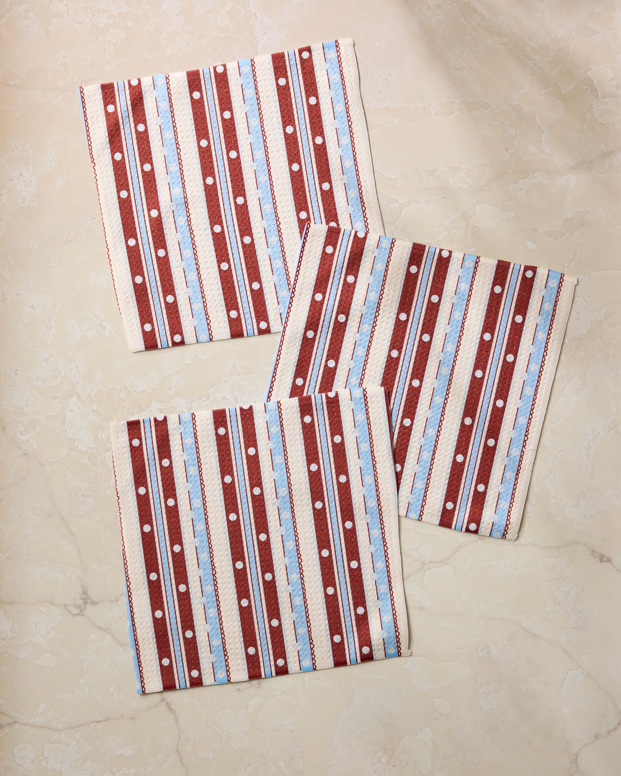 Three folded “Blue Skies Stripes” cloth napkins by Geometry, featuring red, white, and blue stripes and polka dots, are arranged on a beige marble surface—an eco-friendly choice for kitchen towels or dishcloths.