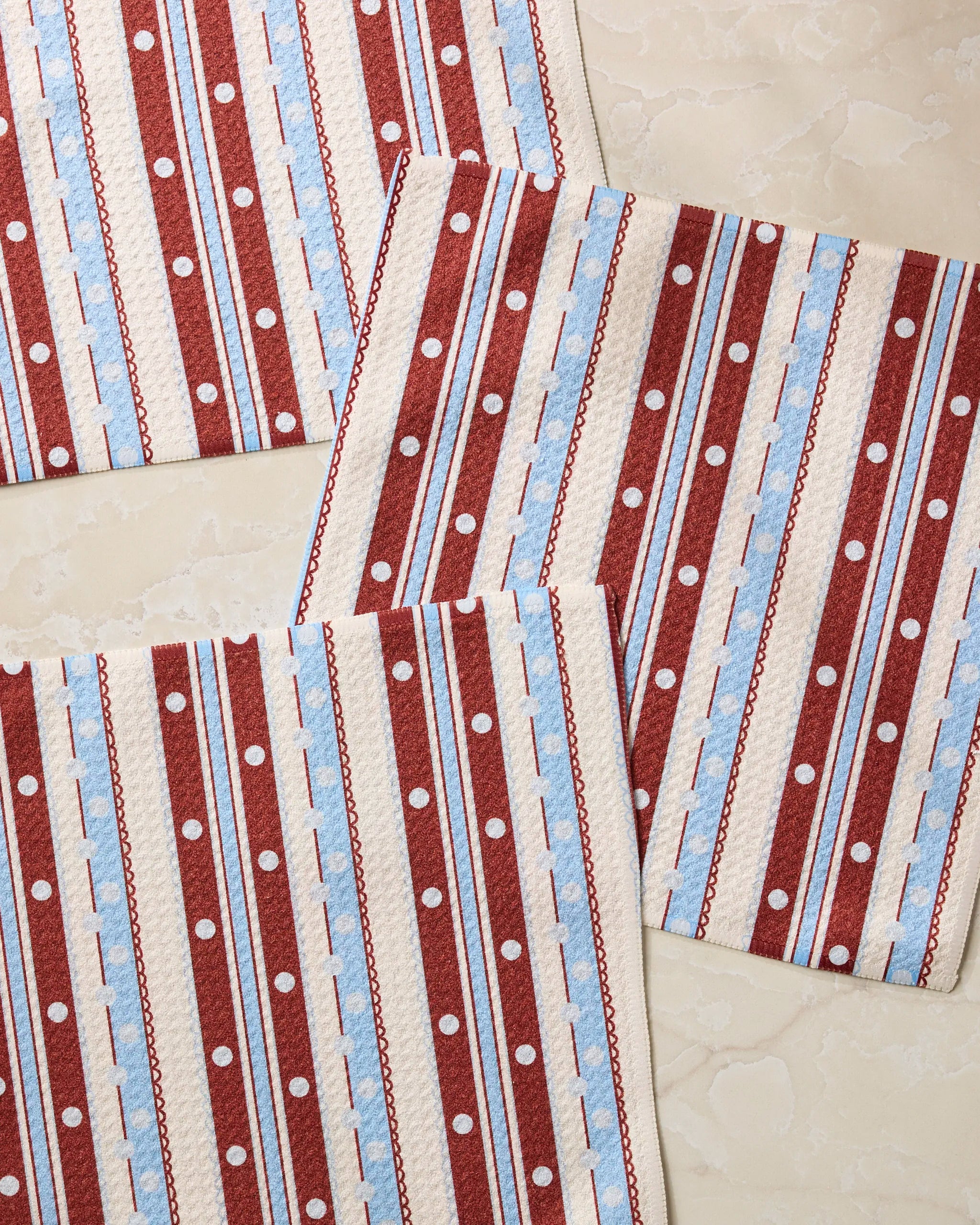 Three "Blue Skies Stripes" cloth napkins by Geometry, featuring red, white, and blue stripes and dots, are arranged with overlapping corners on a marble surface—an eco-friendly addition to your kitchen.