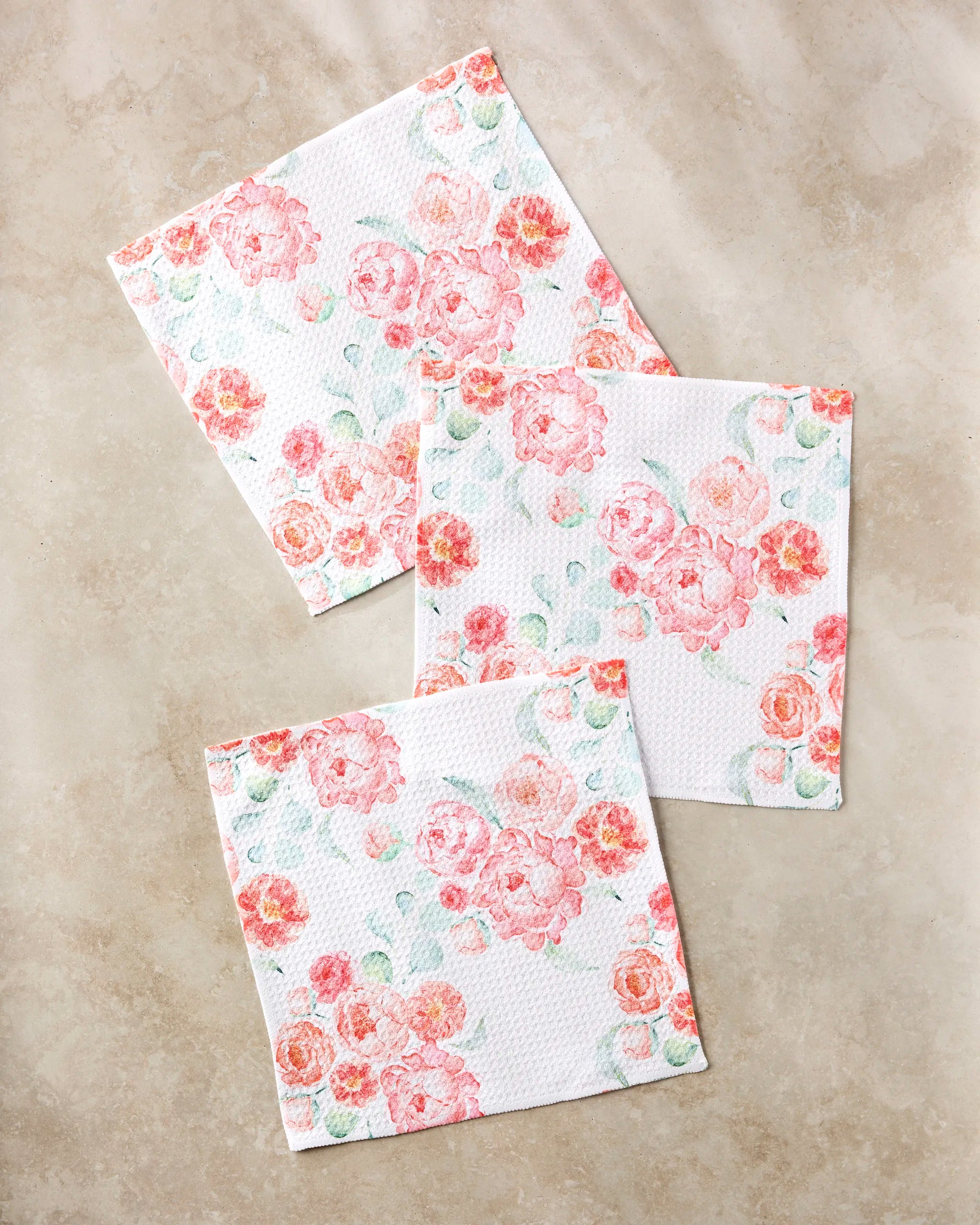 Three square "Blush Florals" napkins by Elena Fay, made from recycled materials, feature a pink and peach floral pattern with delicate green leaves, arranged elegantly on a light beige marble surface.