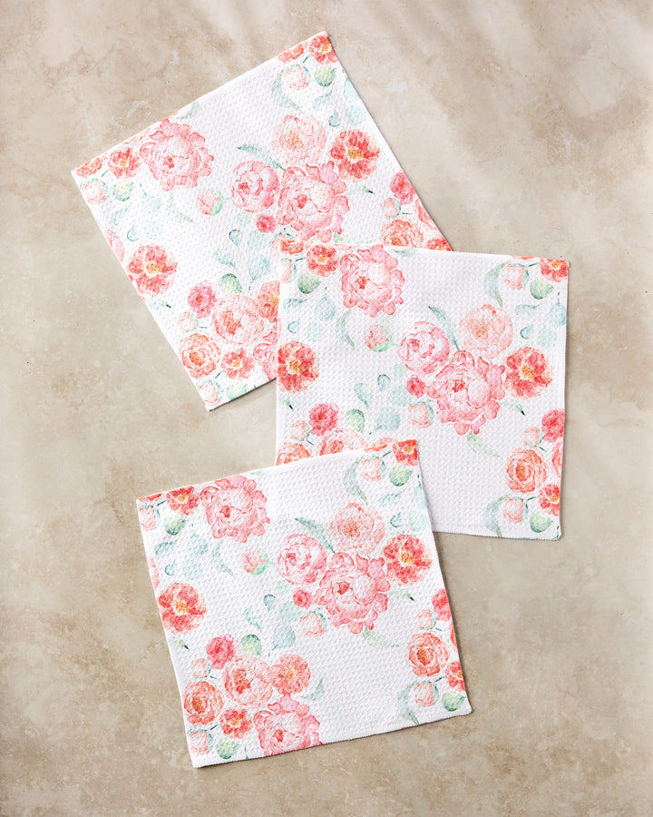 Three square "Blush Florals" napkins by Elena Fay, made from recycled materials, feature a pink and peach floral pattern with delicate green leaves, arranged elegantly on a light beige marble surface.