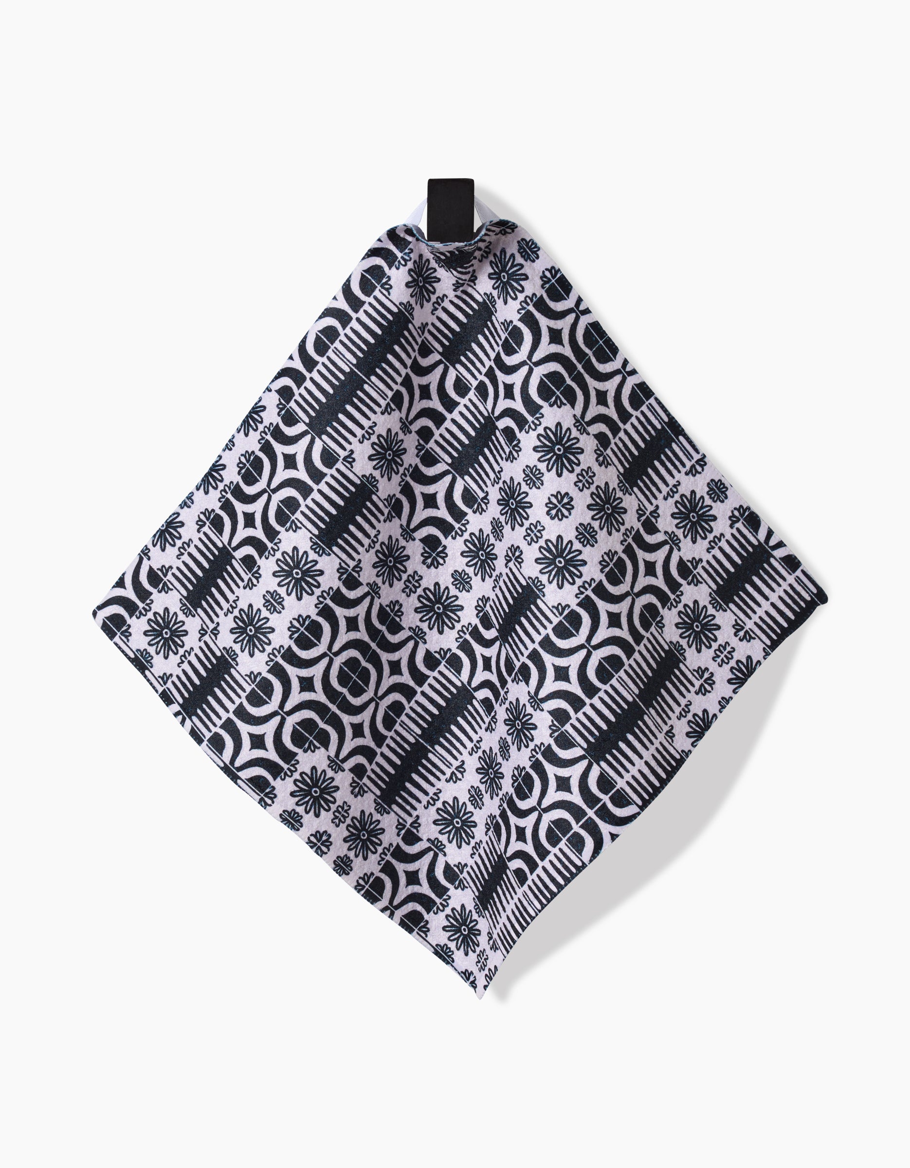 The GEOMETRY Bohemian Mosaic dishcloth, made from Geoweave fabric, features a black and white geometric and floral tile pattern and is shown hanging by a black loop against a white background.