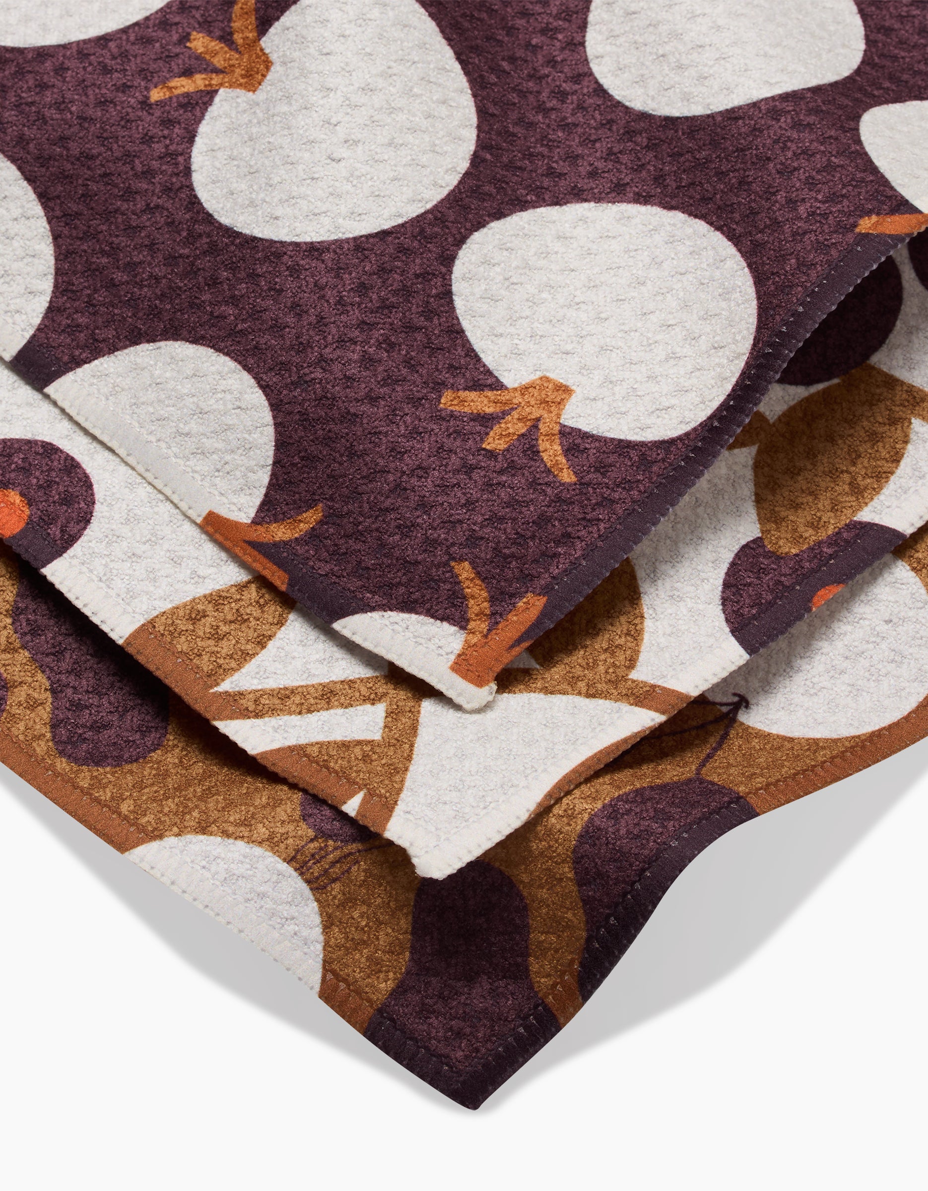 A close-up of Bold Fall dishcloths by Lizzie Clark, folded to show their textured, recycled fabric with large white and brown abstract shapes on a dark maroon background.
